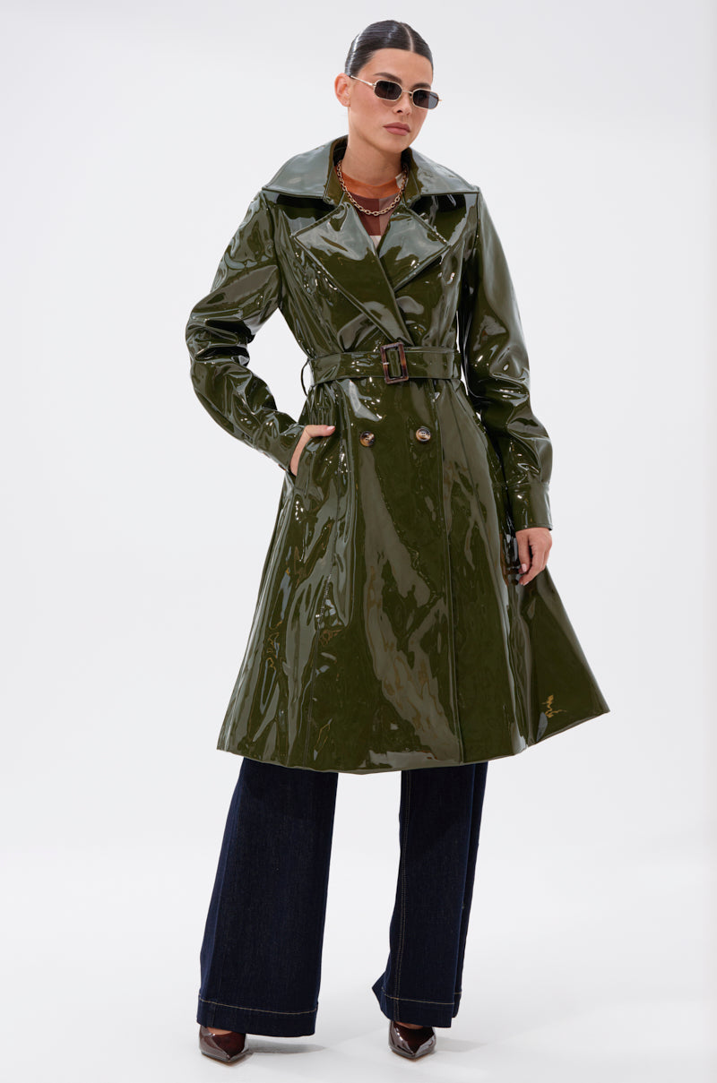 A woman poses confidently against a plain white background wearing the MITICA PATENT TRENCH IN OLIVE, paired with dark wide-leg pants, pointed shoes, sunglasses, and jewelry.