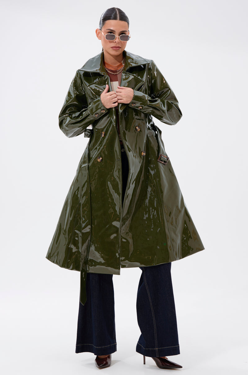 A woman in the MITICA PATENT TRENCH IN OLIVE, sunglasses, dark wide-leg pants, and pointed-toe boots poses confidently against a plain white background.
