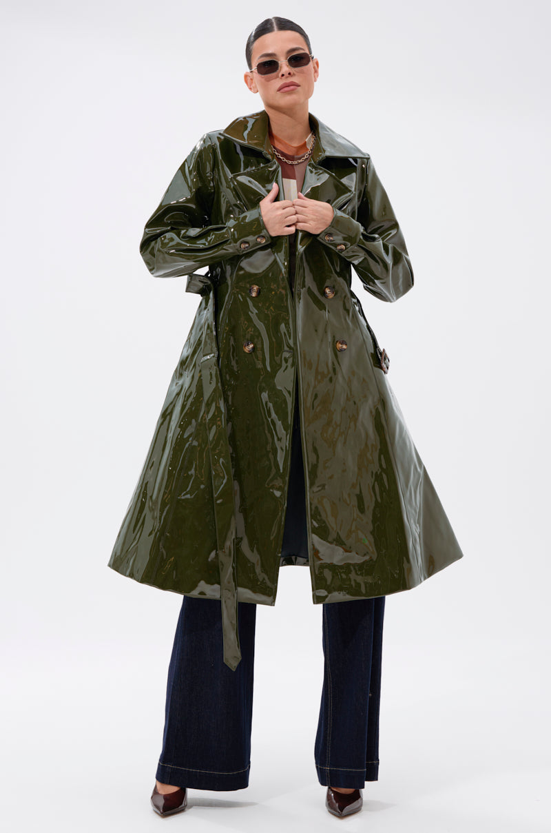 Wearing the MITICA PATENT TRENCH IN OLIVE with a belt, dark flared pants, dark shoes, sunglasses, and hair pulled back, a person stands against a plain white background.