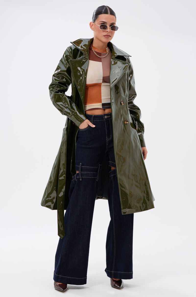 A person in sunglasses, a cropped multicolor top, wide-leg dark denim jeans, and the MITICA PATENT TRENCH IN OLIVE stands confidently against a plain white background.