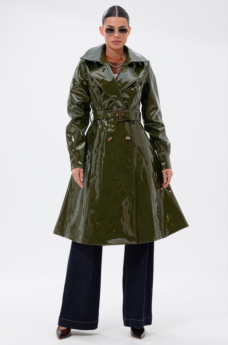A person stands facing forward against a plain white background, wearing the MITICA PATENT TRENCH IN OLIVE, sunglasses, wide-leg dark jeans, and pointed brown shoes.
