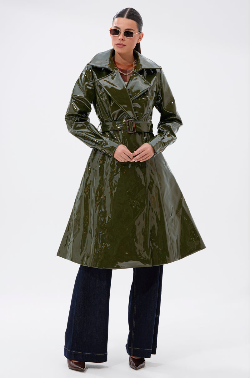 Wearing the MITICA PATENT TRENCH IN OLIVE over wide-leg dark jeans and brown shoes, with sunglasses and hair pulled back, a person stands against a plain white background.