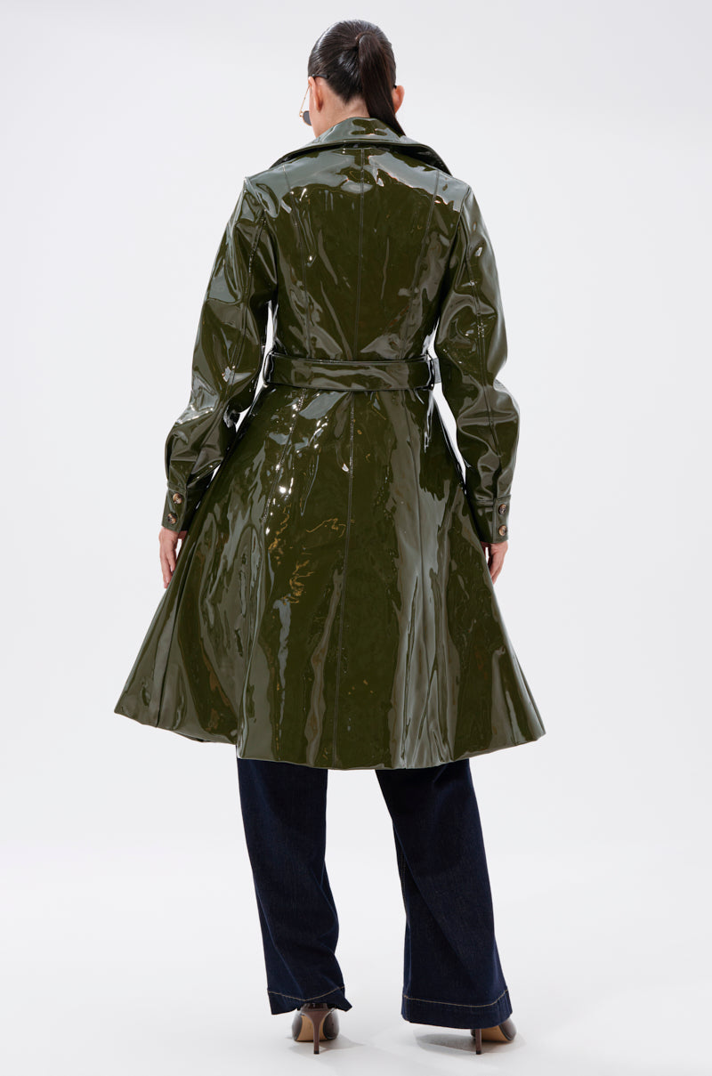 A person faces away wearing the MITICA PATENT TRENCH IN OLIVE, a shiny faux patent leather coat with a belt, paired with dark wide-leg pants and beige shoes. Their hair is tied back, all set against a plain white background.
