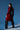 A woman stands against a blue background wearing the shiny red MITICA PATENT TRENCH COAT and black ruffled high-heeled boots, looking over her shoulder with a confident expression.
