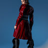 A woman stands against a blue background wearing the shiny red MITICA PATENT TRENCH COAT and black ruffled high-heeled boots, looking over her shoulder with a confident expression.