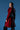 A woman stands confidently against a blue background, looking over her shoulder while wearing the MITICA PATENT TRENCH COAT in shiny red faux leather with a studded belt detail.