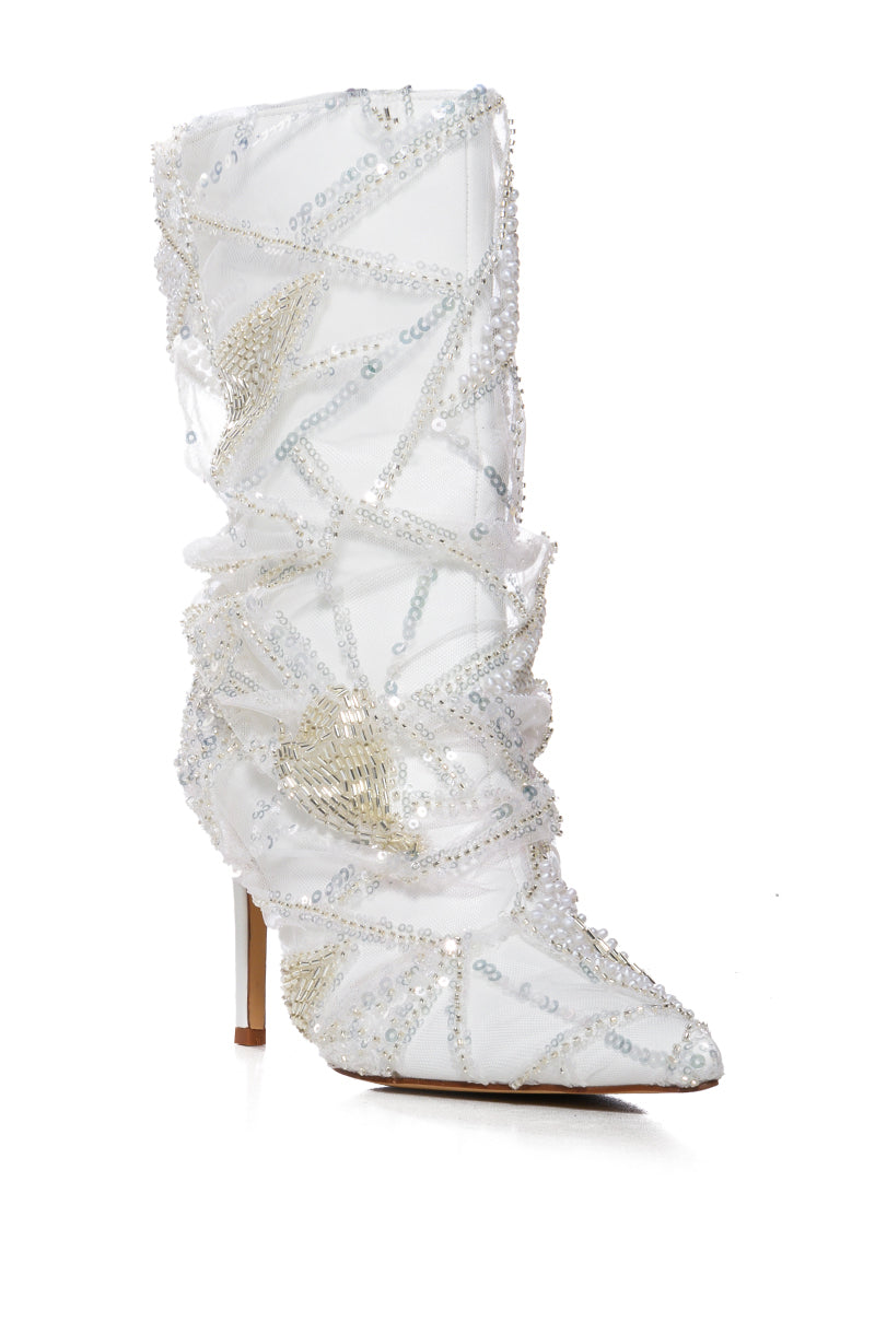 The AZALEA MITSKI WHITE SEQUIN OVERLAY BOOTIE is a white high-heeled boot with a faux leather upper, detailed with swirling beadwork and sequins. It features a pointed toe and stiletto heel against a plain white backdrop.