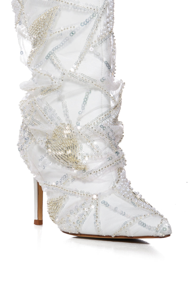 The AZALEA MITSKI WHITE SEQUIN OVERLAY BOOTIE features a white stiletto heel, faux leather upper, and a pointed toe. It is adorned with intricate pearl and sequin embellishments in geometric patterns, enhancing its stylish and luxurious appearance.
