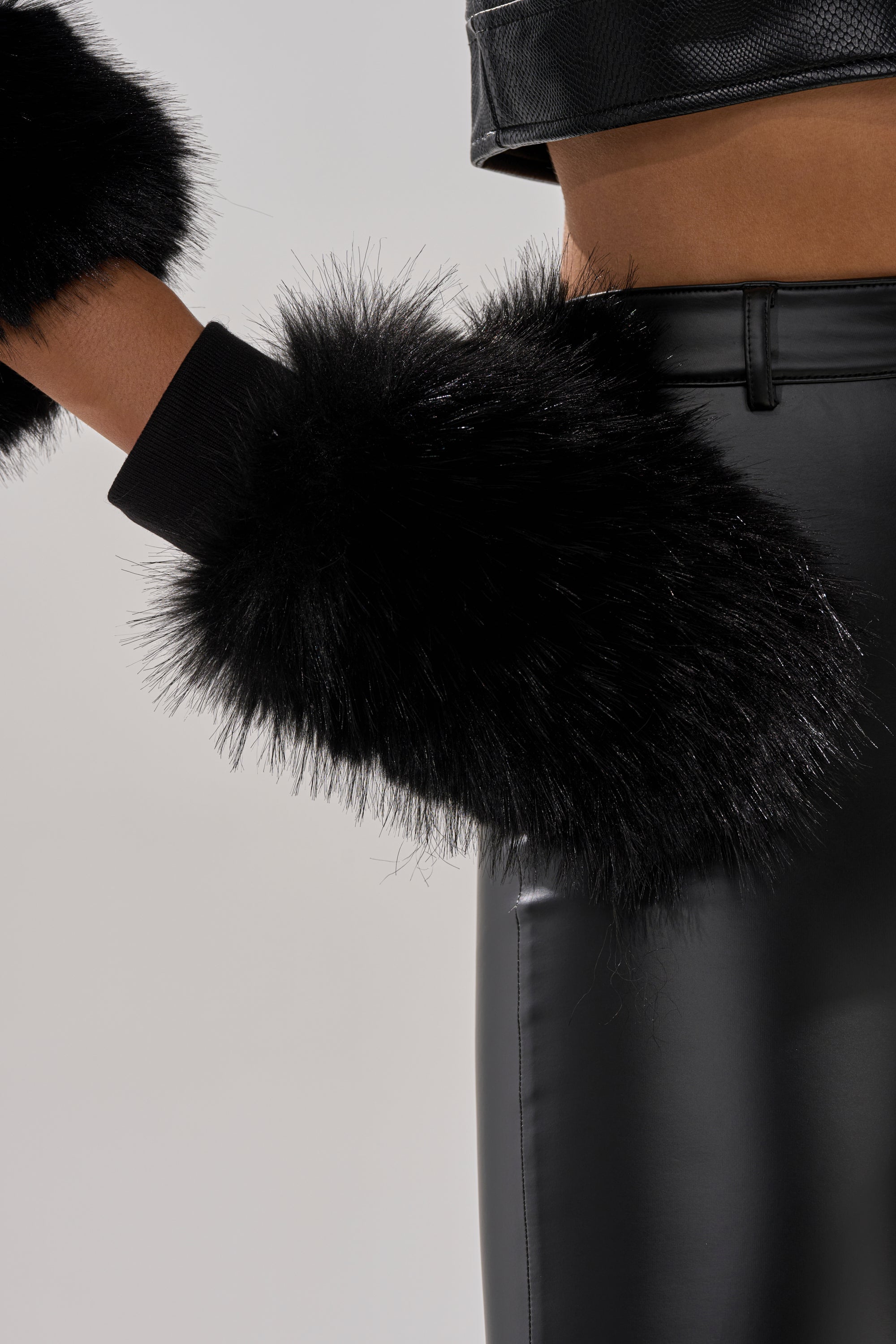 A close-up of a person wearing IN THE NIGHT FAUX FUR MITTENS and black leather pants, with part of a black textured jacket—perfect winter wardrobe inspo against a light, plain background. 0