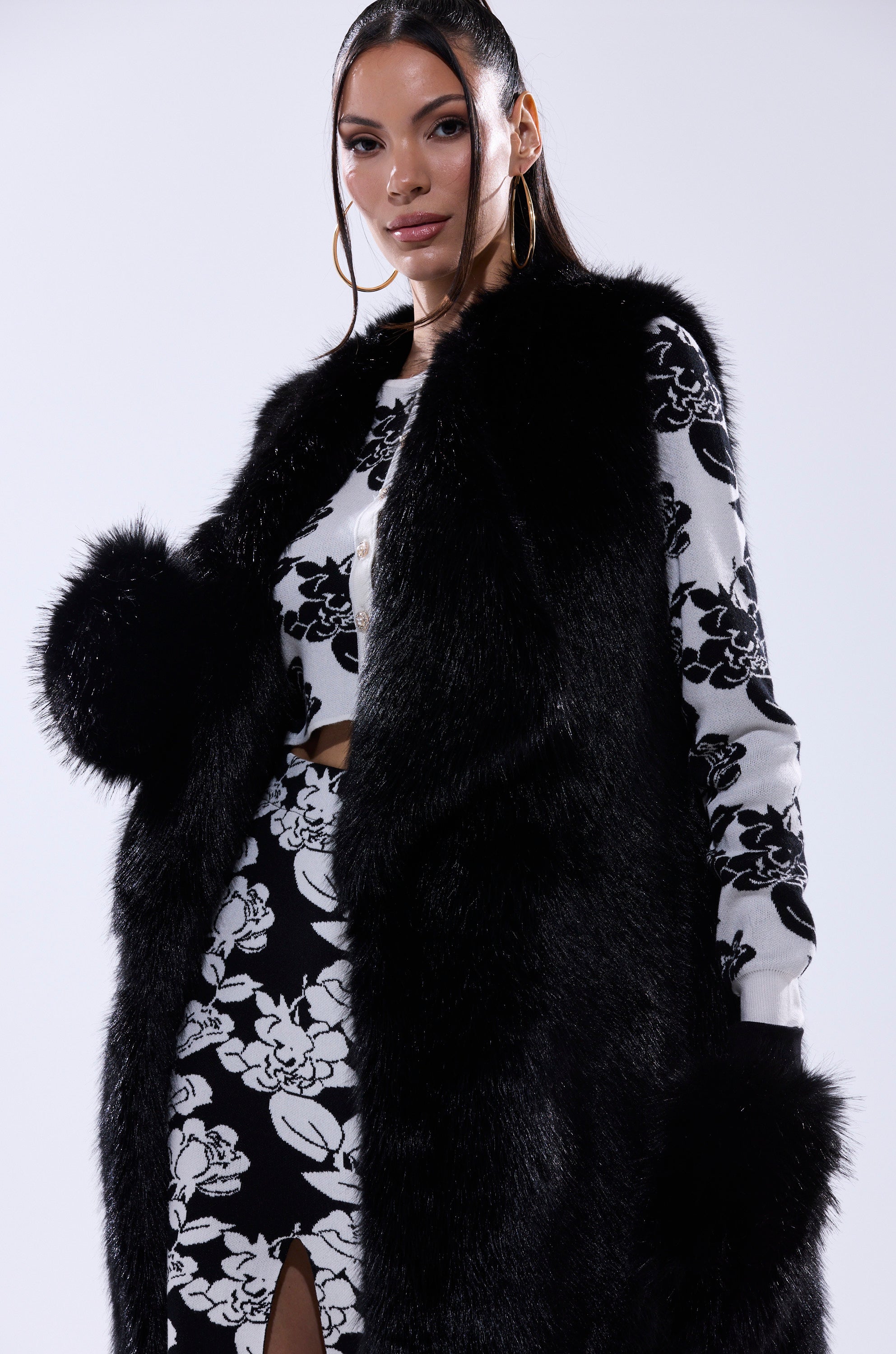 A woman channels confidence with her hair styled back and large hoop earrings, wearing IN THE NIGHT FAUX FUR MITTENS paired with a chic black and white floral ensemble—perfect inspiration for your winter wardrobe. 6