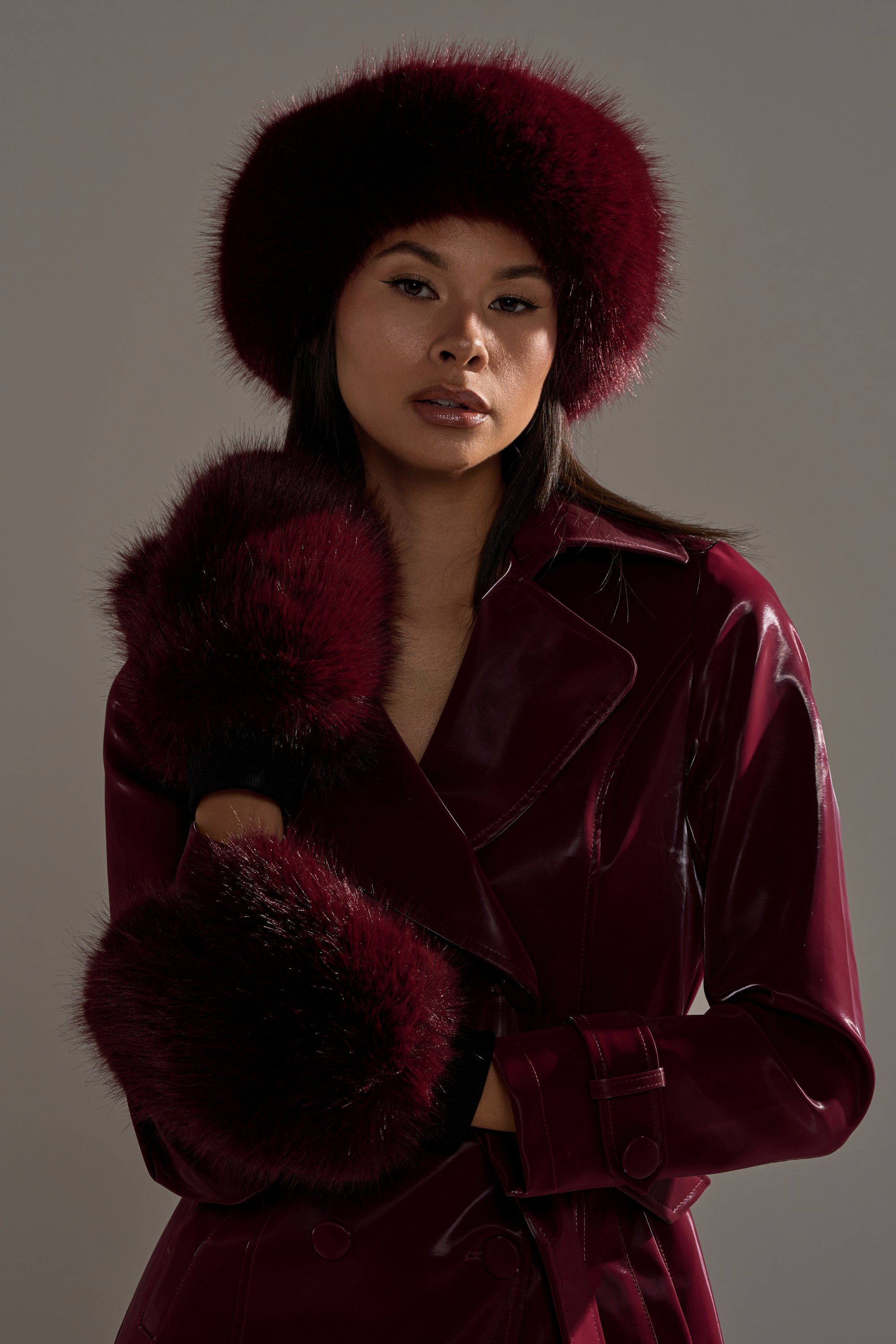 A person confidently poses against a plain gray background, wearing a shiny burgundy trench coat, a matching burgundy fur hat, and ALAIA FAUX FUR MITTENS.