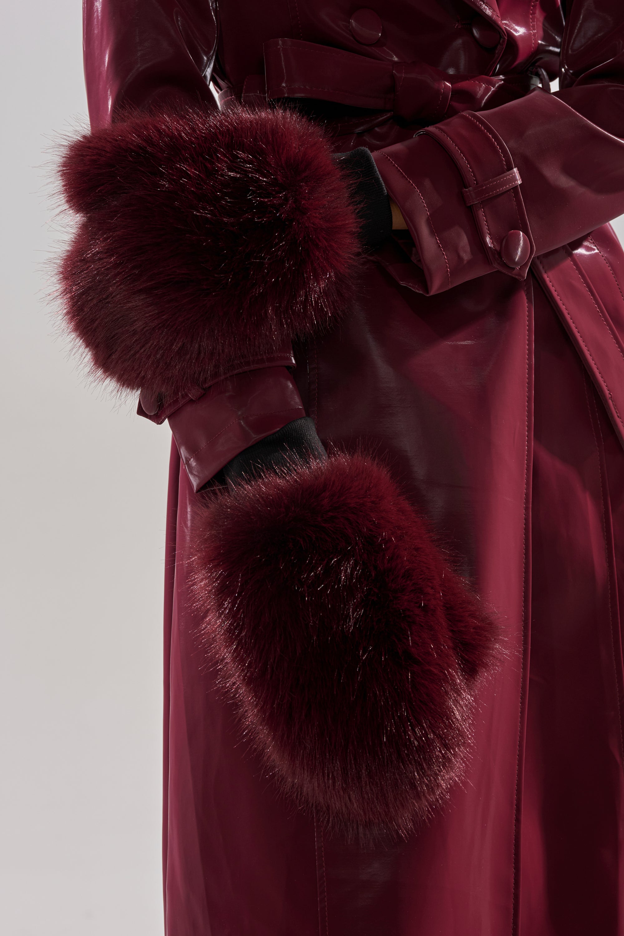 Wearing ALAIA FAUX FUR MITTENS, paired with a glossy burgundy leather coat with fluffy fur cuffs over black gloves, this look is perfect for elevating any winter wardrobe against a plain light background.