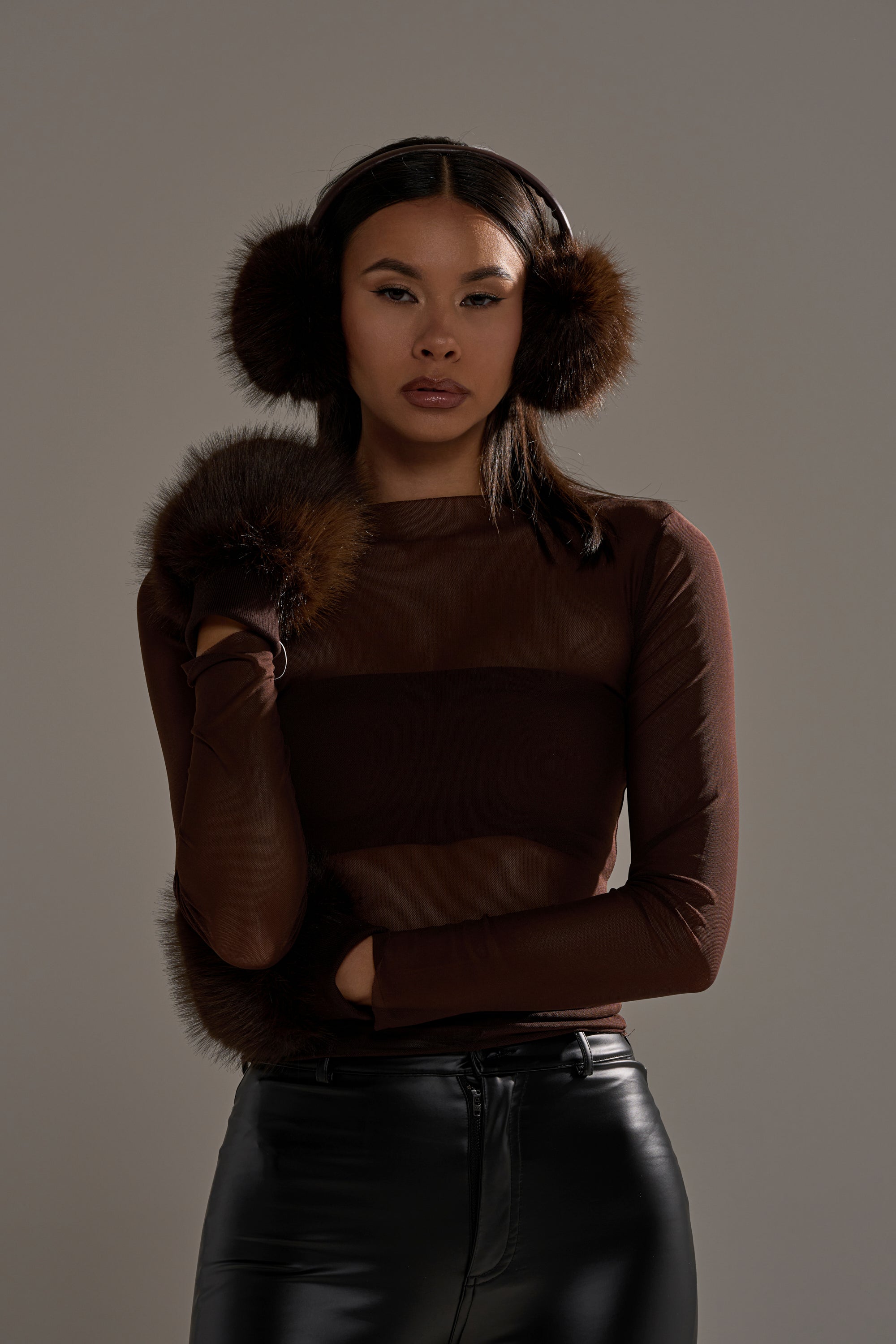 A woman in brown fur earmuffs, a sheer brown long-sleeve top, black leather pants, and DARK CHOCOLATE FAUX FUR MITTENS stands against a plain gray background, gazing at the camera with a neutral expression. 0