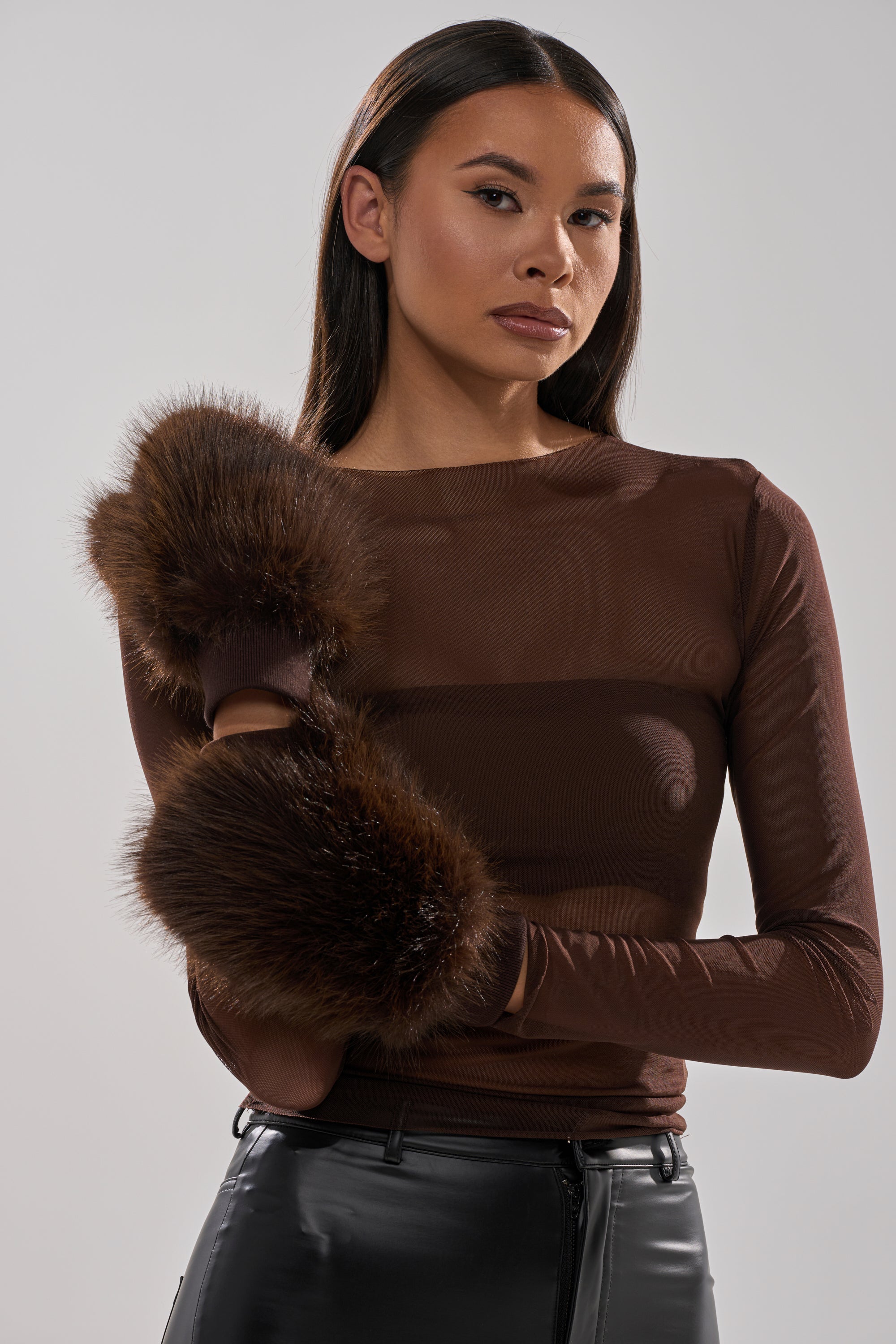 A woman with long dark hair wears DARK CHOCOLATE FAUX FUR MITTENS, a sheer brown top, and black leather pants—perfect additions to your winter wardrobe. She stands before a light gray background, confidently facing the camera with her arms crossed. 2