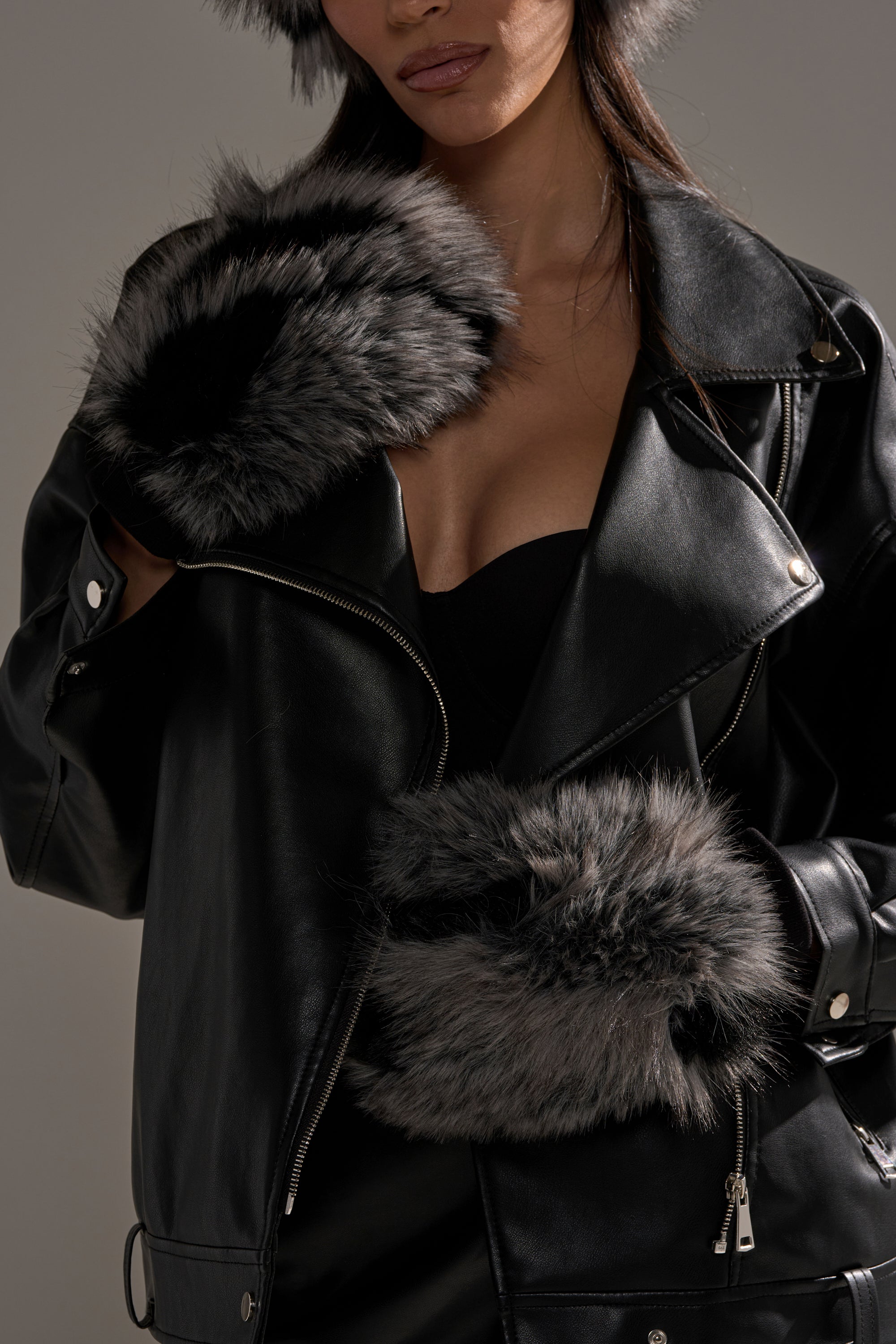 A woman wears a black leather jacket, black top, and plush gray ARLO FAUX FUR MITTENS with a matching hat. The image is cropped at the nose, drawing attention to her chic winter style and standout accessories. 1
