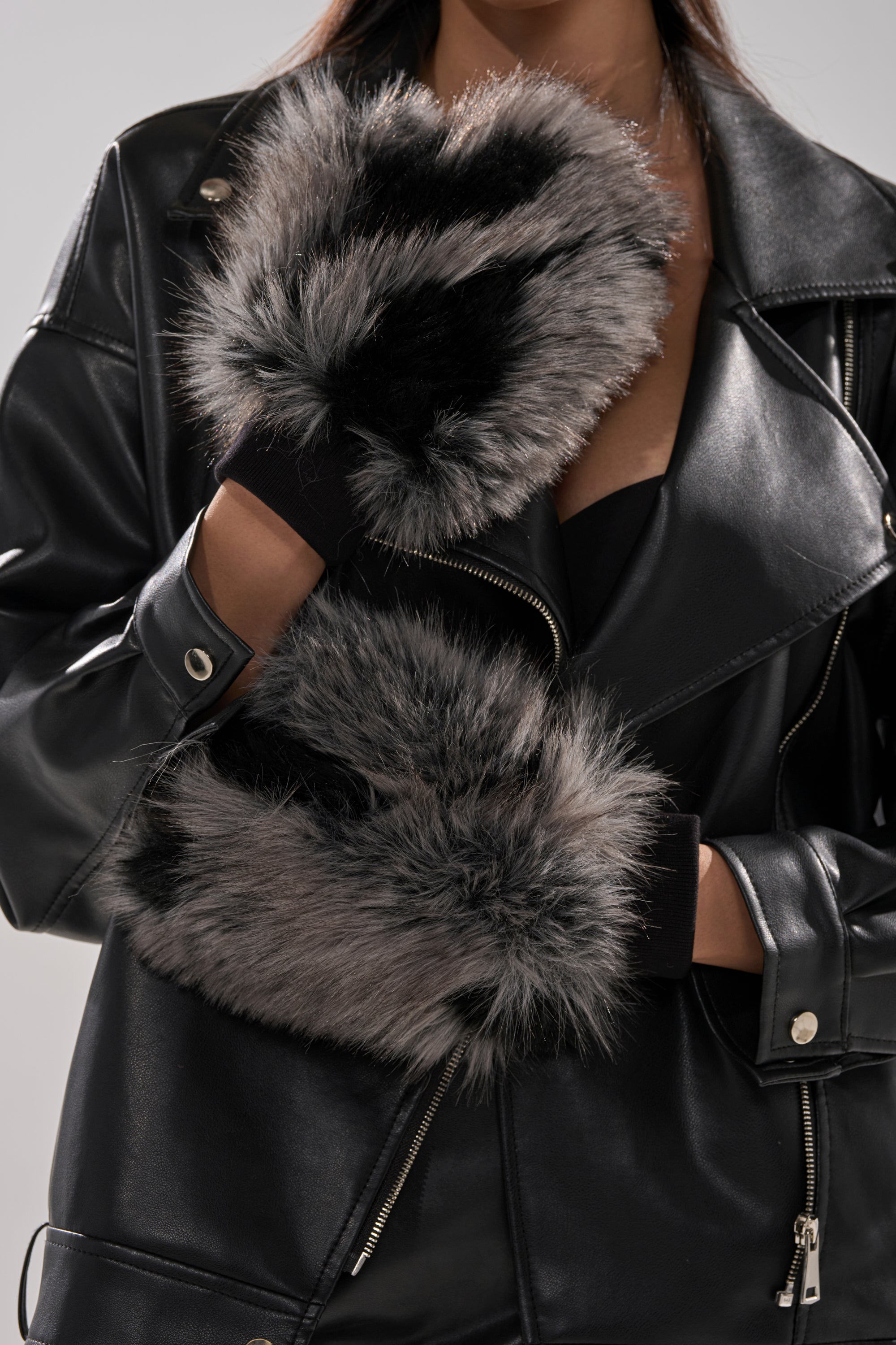 A person wearing a black leather jacket and ARLO FAUX FUR MITTENS—plush mittens with gray and black faux fur accents—holds their hands near their chest, adding cozy style to any winter wardrobe. 3