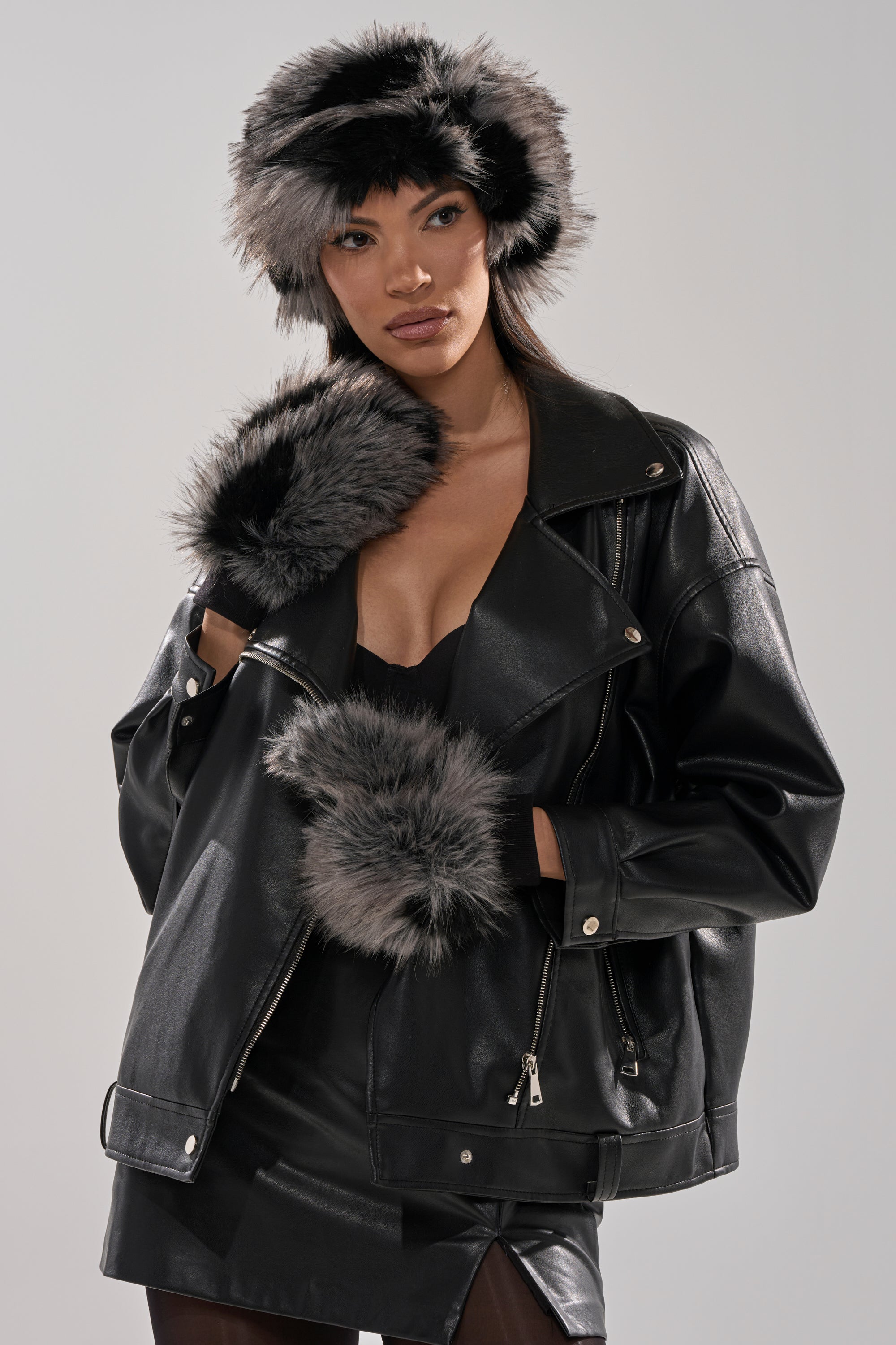 Wearing a black leather jacket, skirt, and top, with matching gray-and-black ARLO FAUX FUR MITTENS plus a plush faux fur hat and scarf, she stands against a plain background—an elegant winter wardrobe look. 4