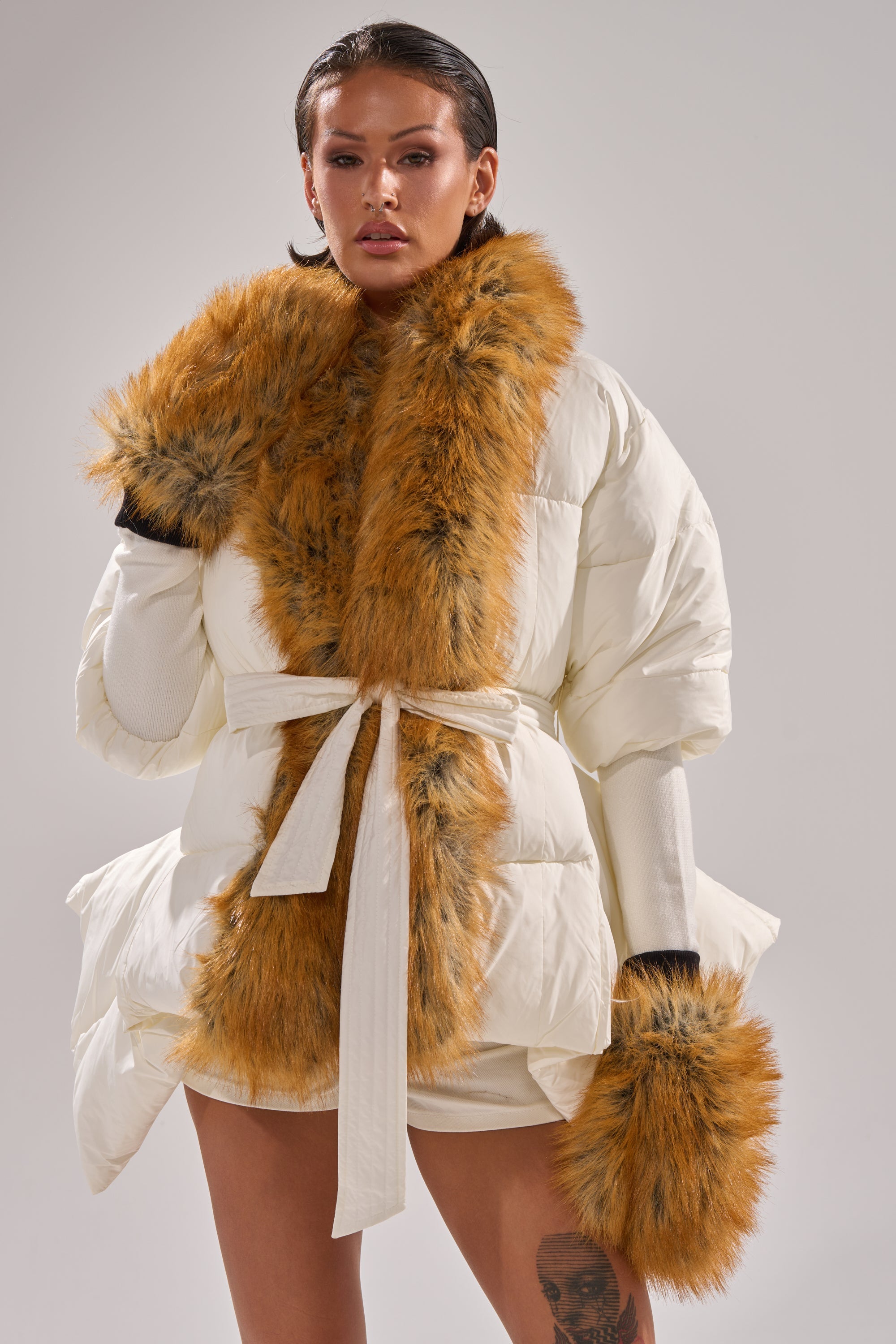 A woman in a white belted puffer coat with plush orange trim and matching REALLY LIKE THAT FAUX FUR MITTENS stands against a plain light background, her slicked-back hair and visible leg tattoo elevating any winter look. 6