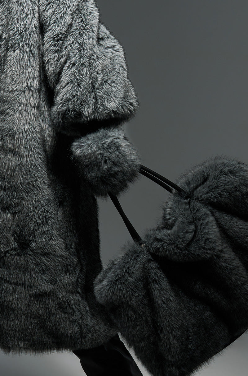 A person wearing a thick gray Tsunami Faux Fur coat holds a pair of matching furry Tsunami Faux Fur Mittens. The image is styled with moody lighting, highlighting the fur's texture against a neutral gray background, capturing the essence of AKIRA Label’s sophisticated design.
