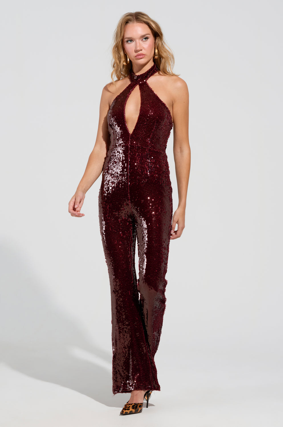 A person radiates confidence in the ON REPEAT SEQUIN JUMPSUIT, a sleeveless, curve-hugging piece adorned with a halter neckline and keyhole cutout, flawlessly complemented by leopard-print high heels. This party-ready ensemble is showcased against a plain, well-lit background.