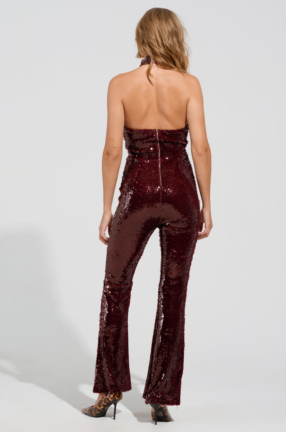A woman with long hair stands with her back to the camera in the ON REPEAT SEQUIN JUMPSUIT, a strapless and curve-hugging burgundy ensemble adorned with sequins, paired gracefully with leopard-print heels. The party-ready jumpsuit showcases a zipper detail down the back against a plain white background.