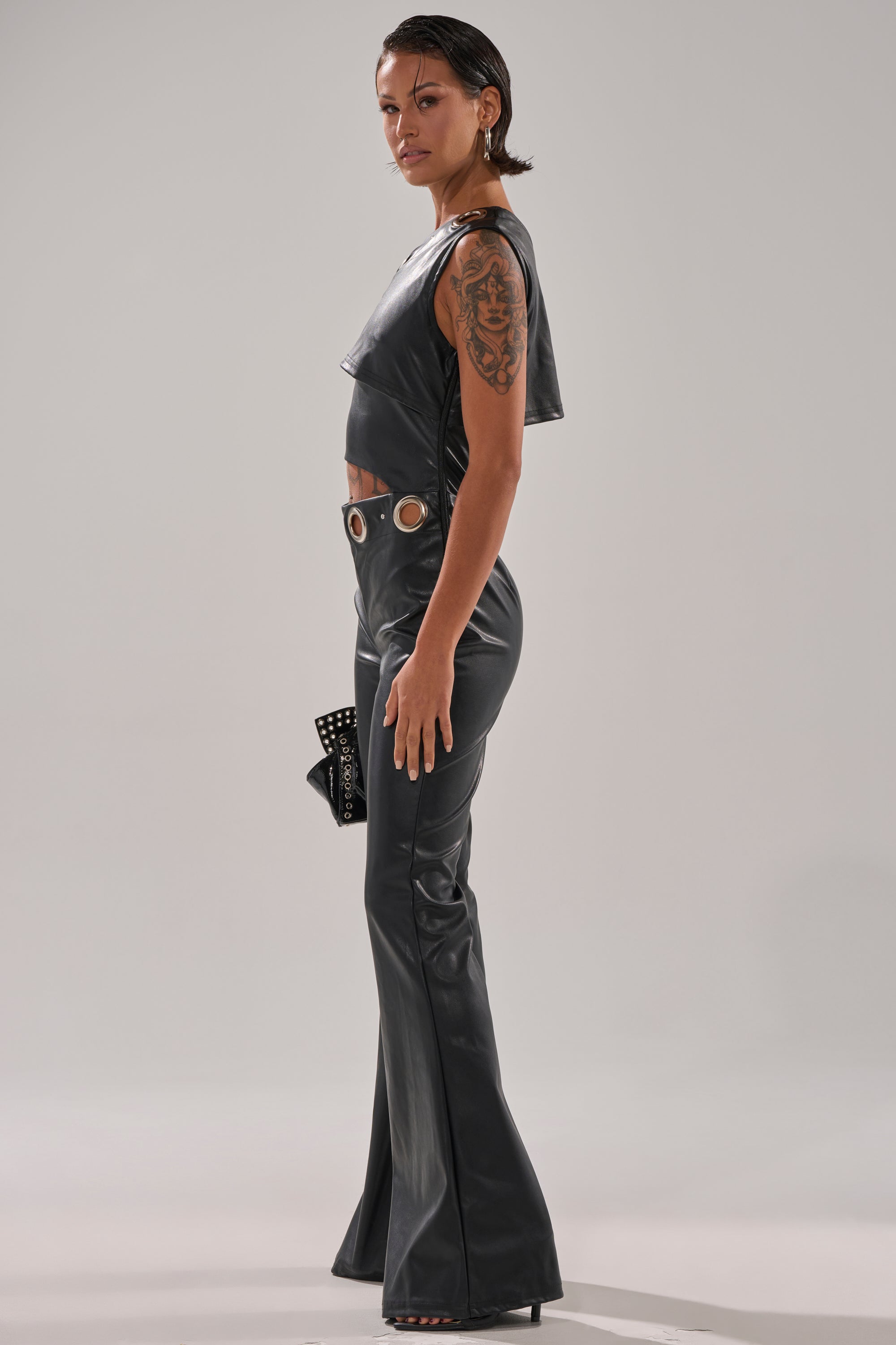 A person with wet-look hair and a large arm tattoo stands sideways, wearing the NO ONE LIKE ME PU JUMPSUIT WITH GROMMETS—featuring shiny black faux leather, flared legs, a grommet belt—and holds a studded clutch bag.