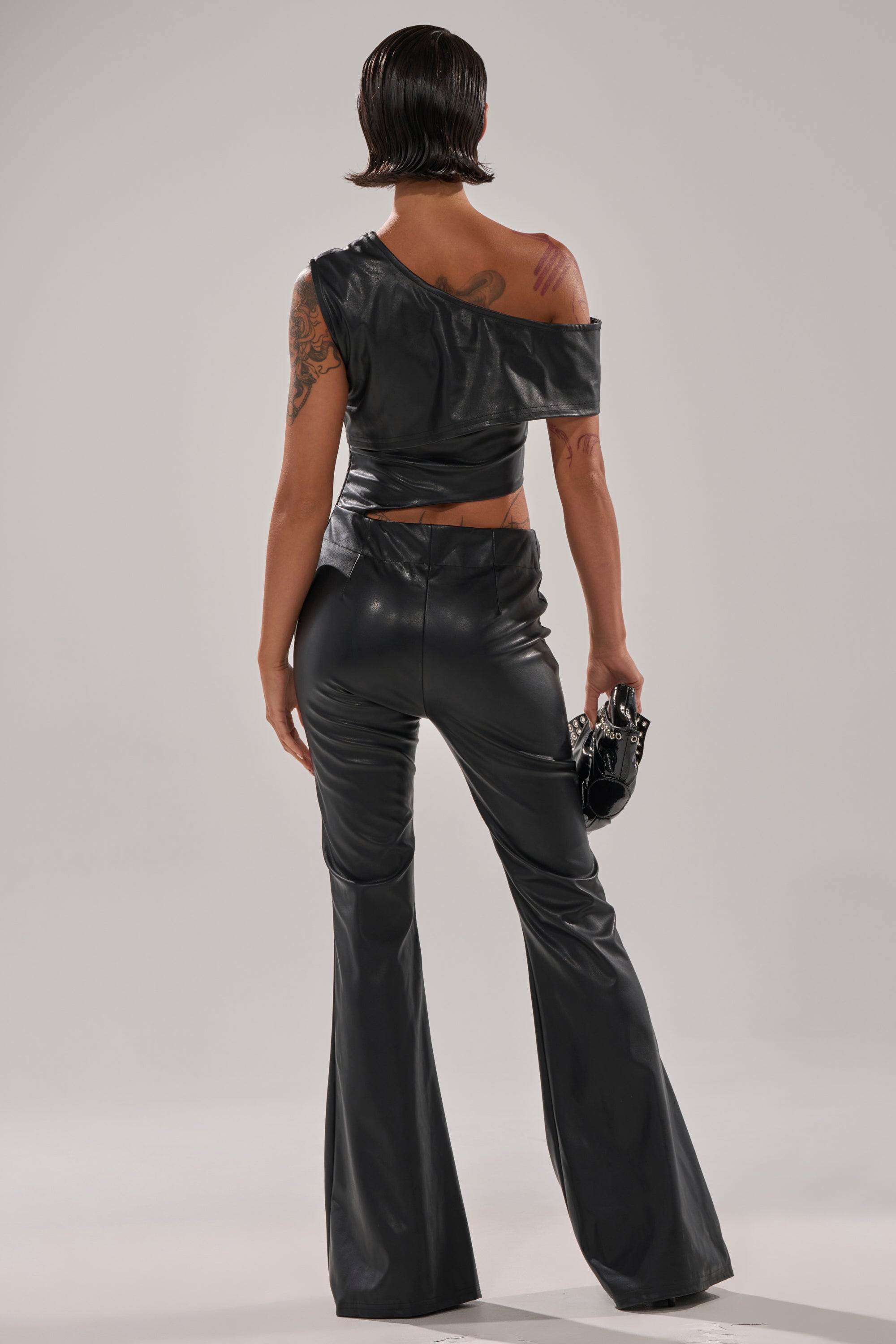 A person with short dark hair and visible tattoos wears the NO ONE LIKE ME PU JUMPSUIT WITH GROMMETS, which is fitted, one-shoulder, and features flared legs. They stand facing away, holding a small black purse.