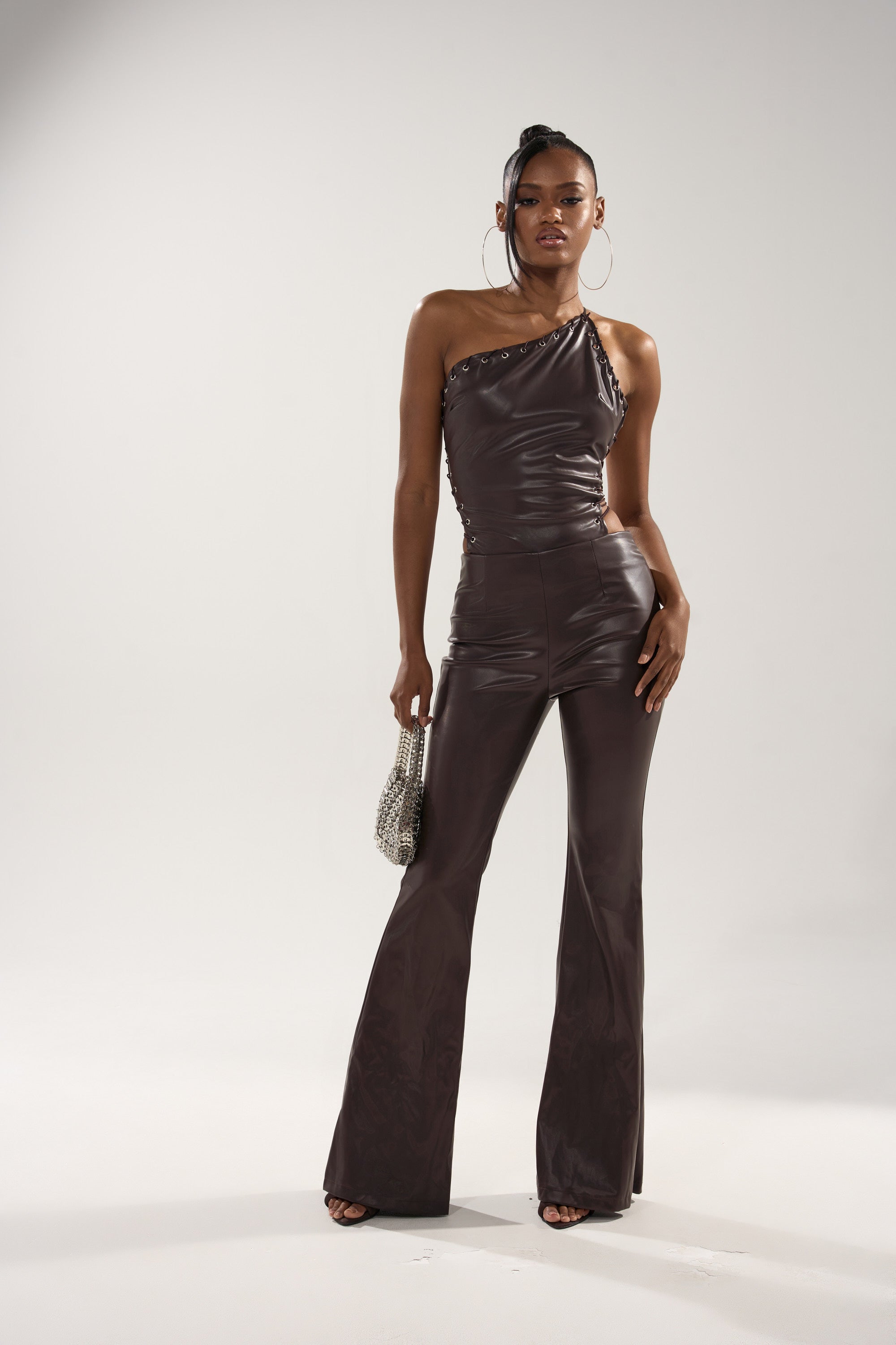 A woman stands confidently in the LEMME SHOW YOU WHAT I GOT PU JUMPSUIT with flared pants, holding a small silver handbag. She wears large hoop earrings and her hair is styled in a bun.