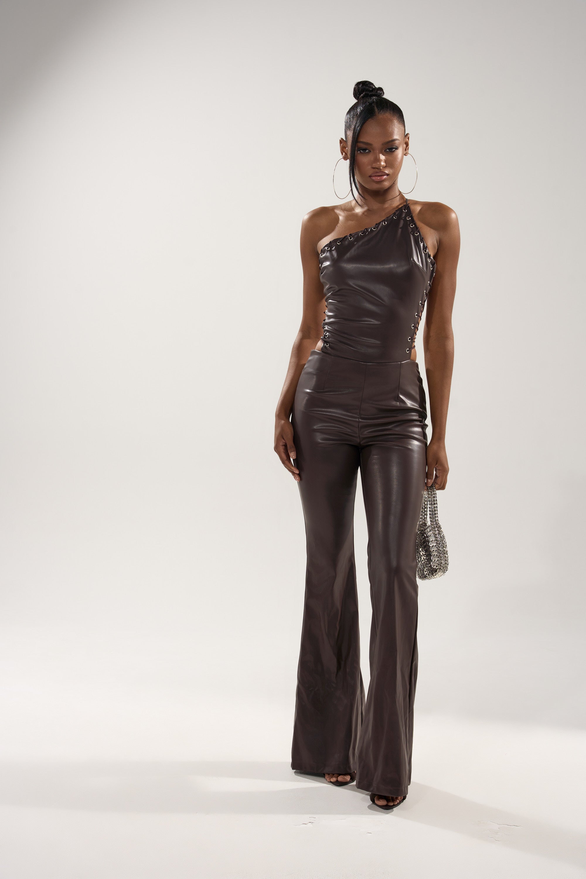 A woman models the LEMME SHOW YOU WHAT I GOT PU JUMPSUIT in chocolate brown, featuring a fitted, one-shoulder design. She accessorizes with a small silver beaded purse and large hoop earrings against a plain background.
