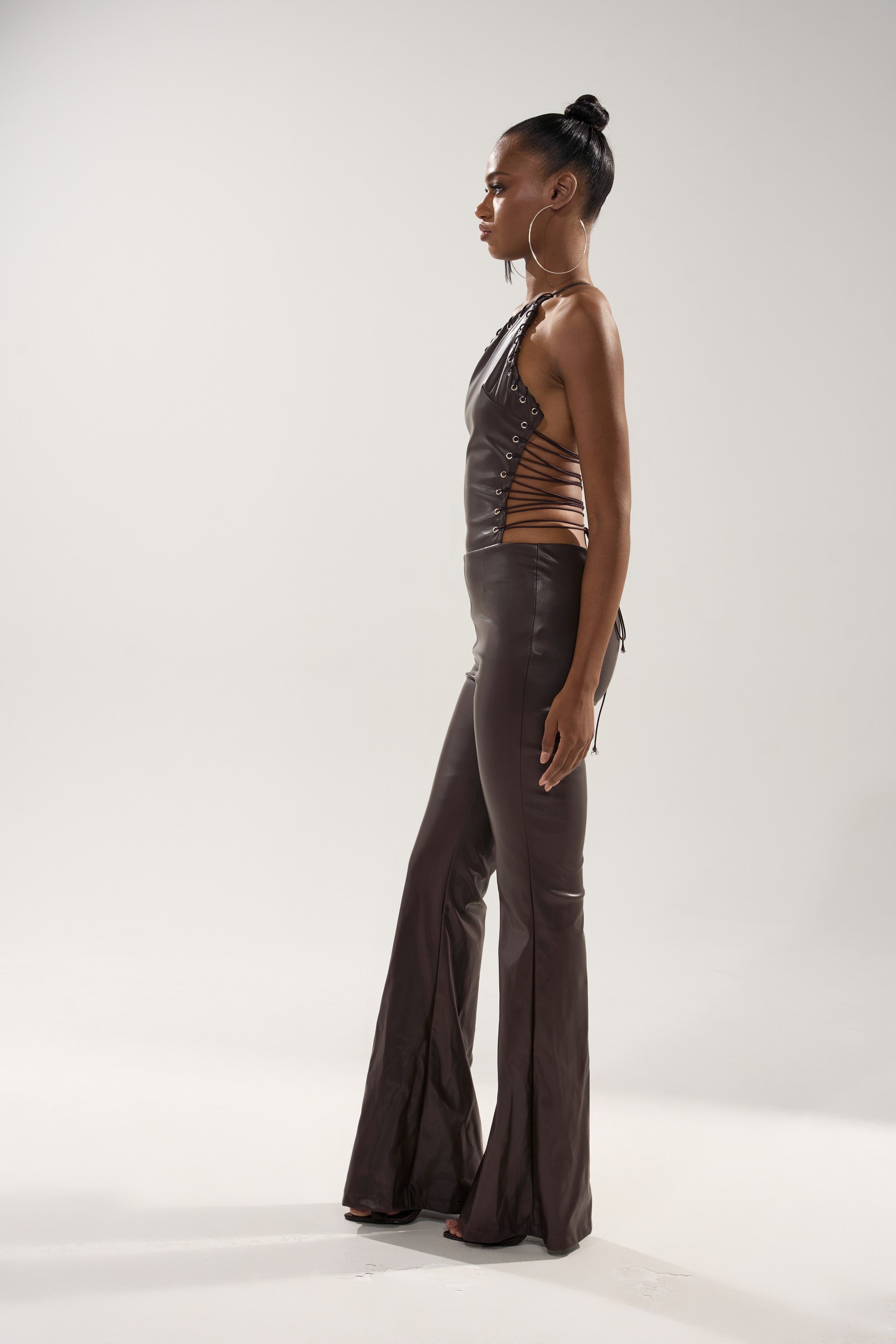 A woman stands in profile against a plain background, wearing the LEMME SHOW YOU WHAT I GOT PU JUMPSUIT. Her hair is in a neat bun, accessorized with large hoop earrings and heeled shoes.