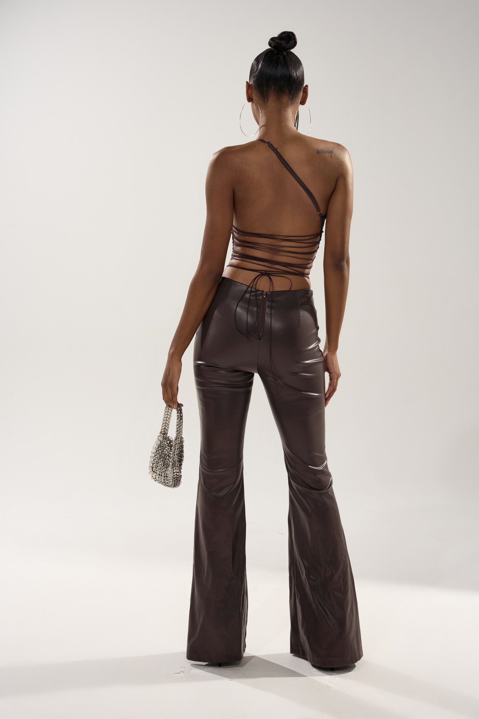 A woman with a neat bun faces away, wearing the LEMME SHOW YOU WHAT I GOT PU JUMPSUIT—chocolate brown, open back, high-waisted flared pants—and holding a small beaded handbag. The background is plain and light-colored.