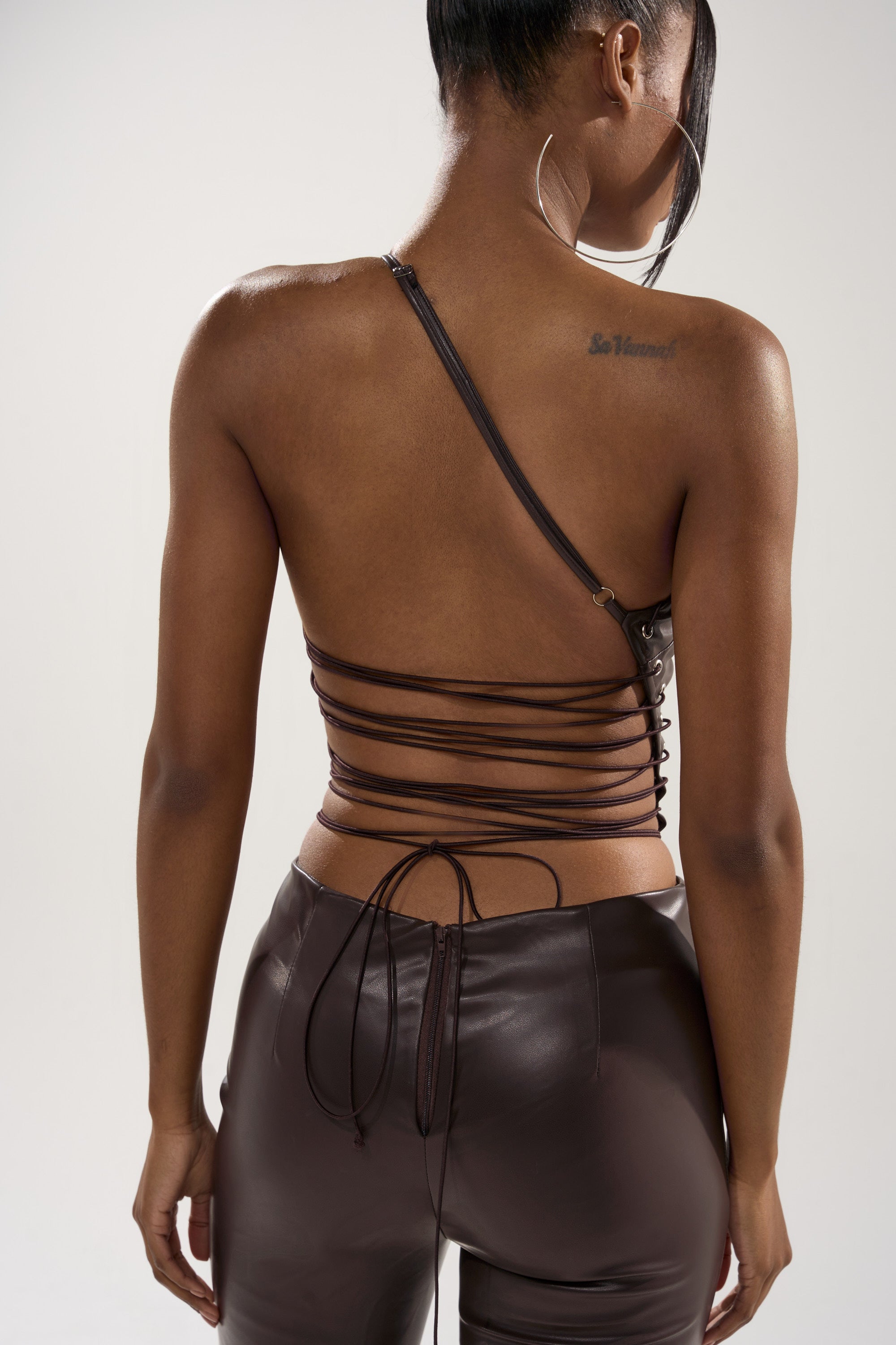 A woman stands with her back to the camera, wearing the LEMME SHOW YOU WHAT I GOT PU JUMPSUIT—a chocolate brown lace-up piece with crisscross straps and faux leather pants. Her hair is tied back, revealing a tattoo on her upper right shoulder.