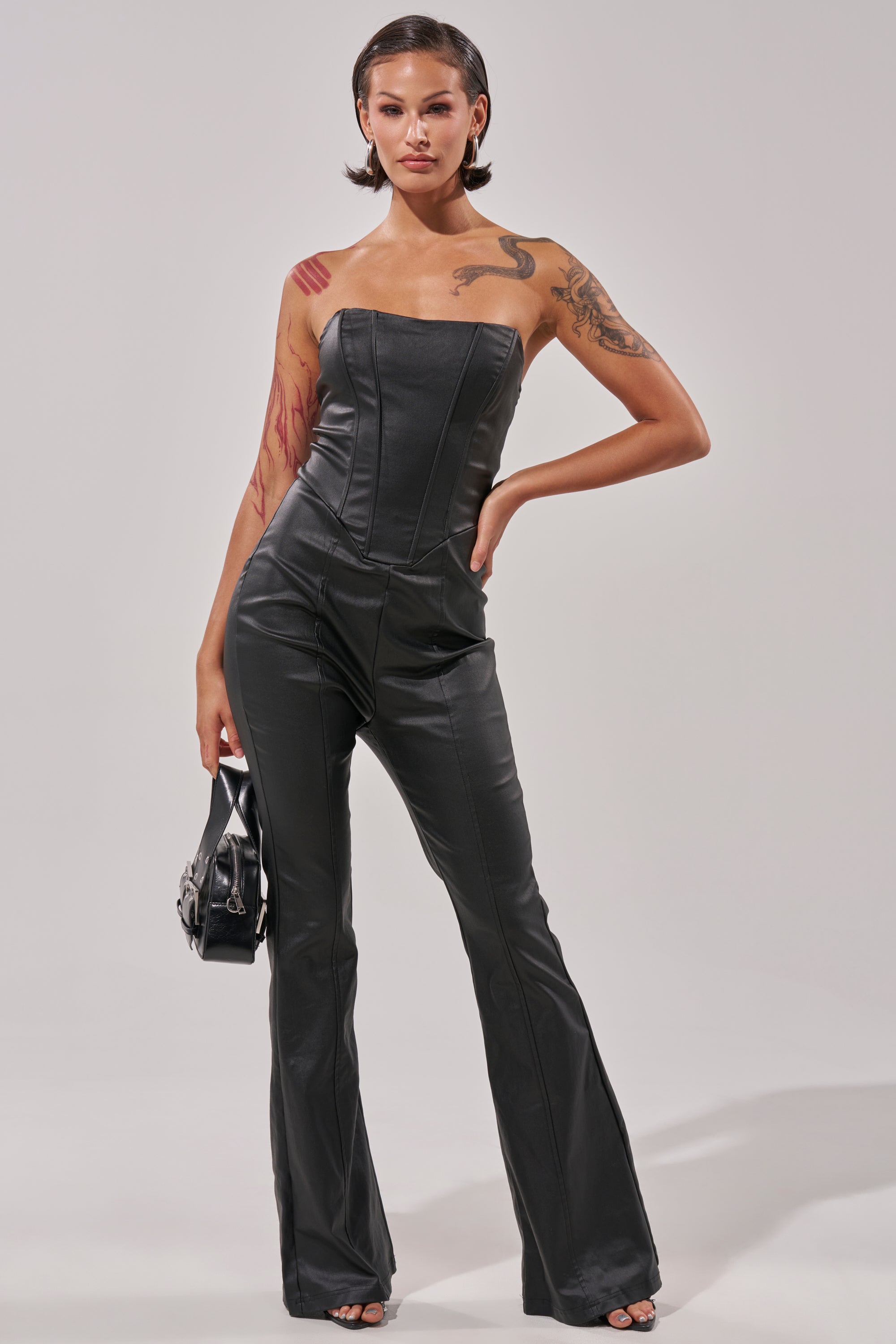 A person with short dark hair models the HOT GIRL PU JUMPSUIT, paired with a small black handbag. Tattoos accent their arm and shoulder as they stand confidently before a plain light background.