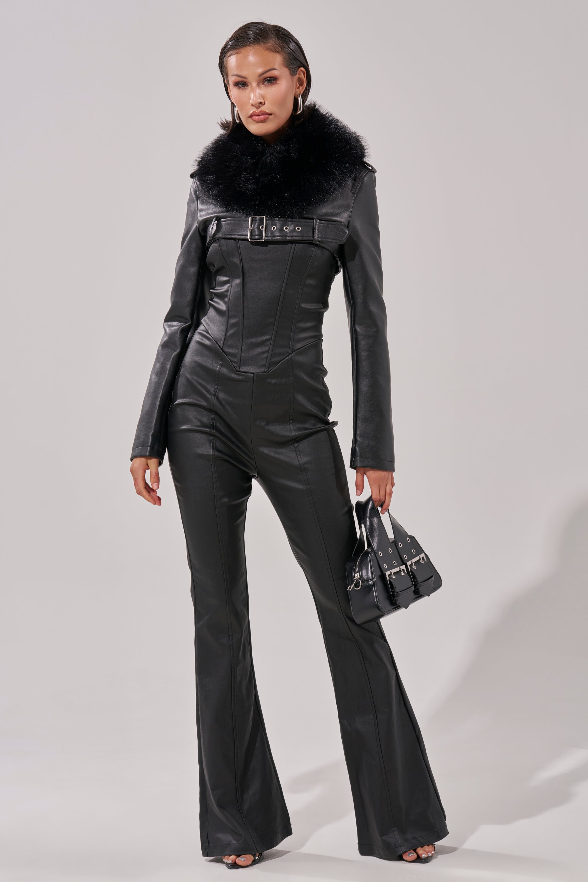 A woman wears the HOT GIRL PU JUMPSUIT, a fitted black faux leather piece with a wide faux fur collar, flared pants, and long sleeves. She accessorizes with hoop earrings, a small black handbag, and slicked-back hair.