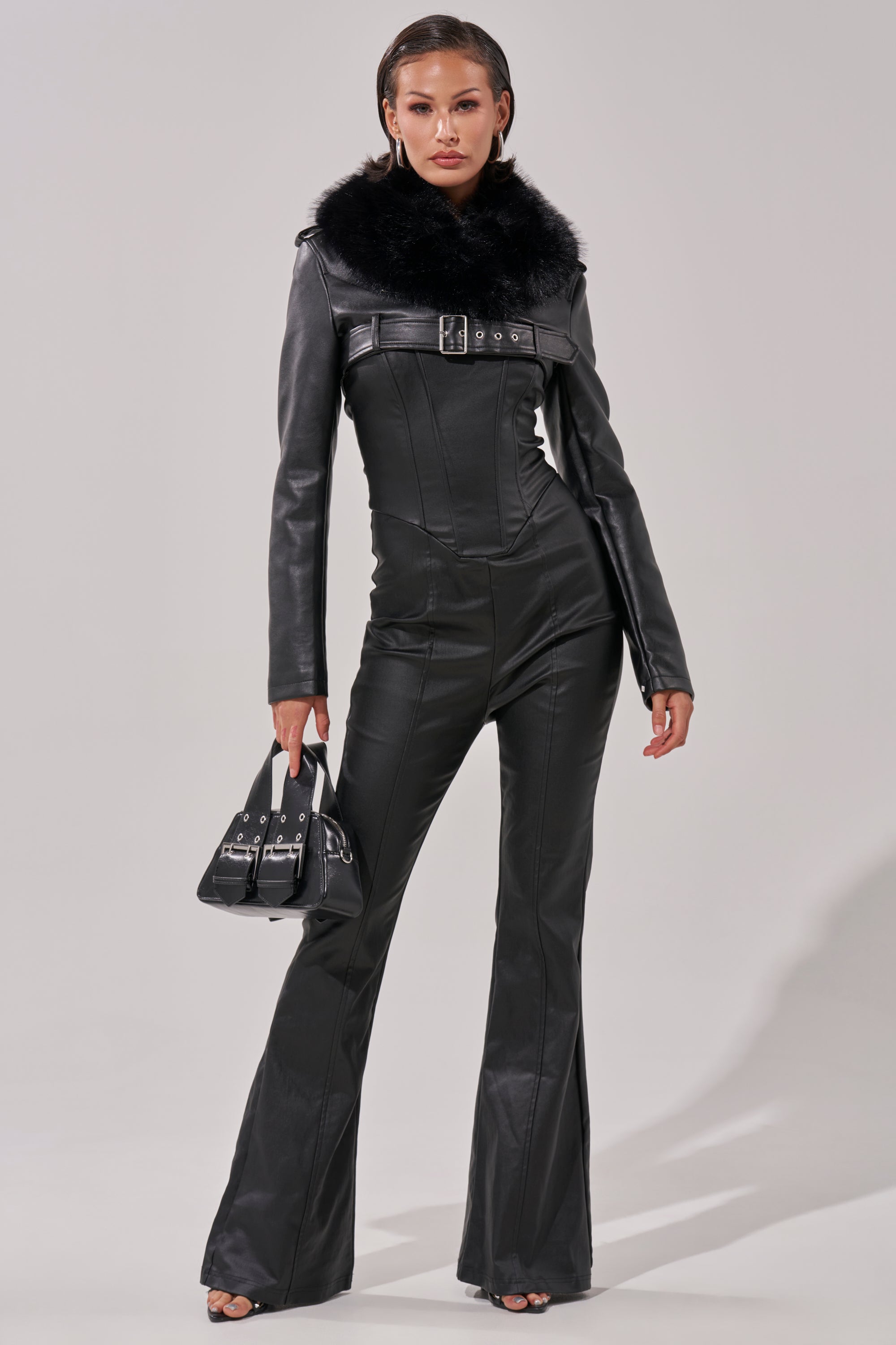 A woman in the HOT GIRL PU JUMPSUIT—black faux leather with flared legs, a belted waist, and a statement faux fur collar—holds a small black handbag, confidently posing against a light background.