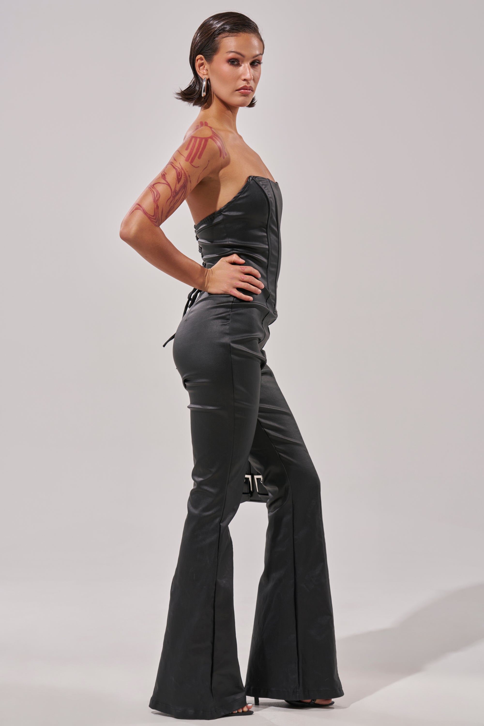 A person with short dark hair and tattoos on their upper arm poses confidently in the HOT GIRL PU JUMPSUIT and heels, standing sideways with one hand on their hip.