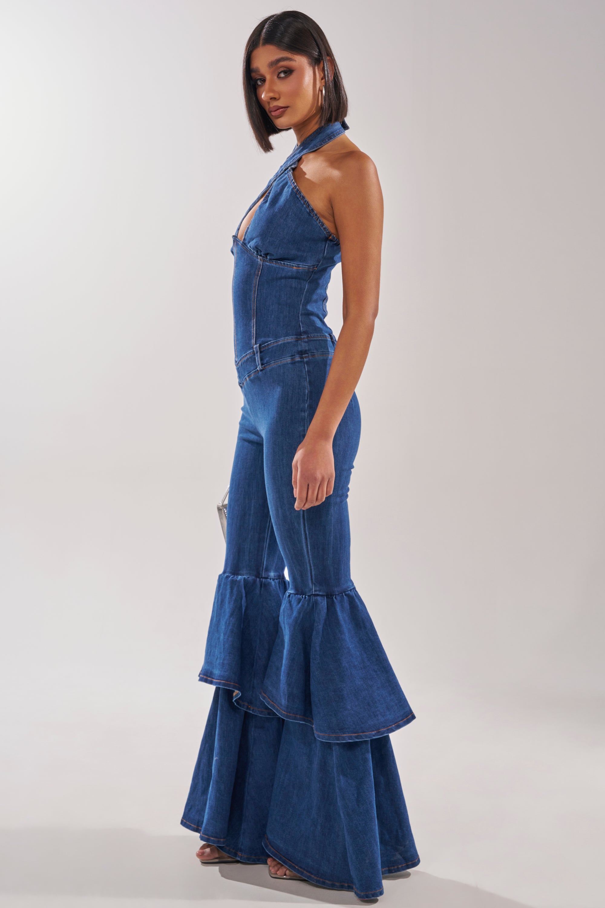 A woman models the RODEO REBEL DENIM JUMPSUIT, a sleeveless halter style with a fitted bodice and tiered ruffle flared legs, channeling retro vibes in a minimalist studio. 3