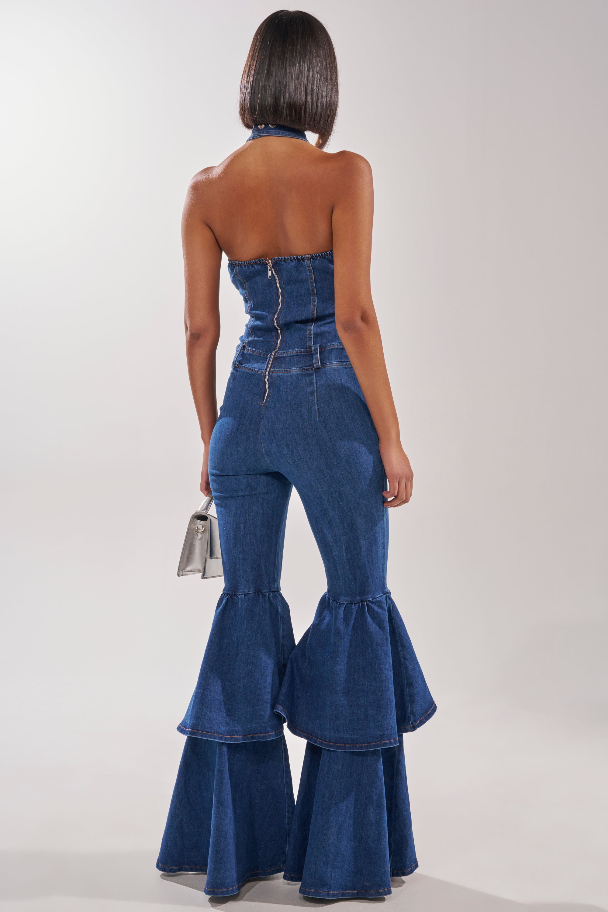 A woman with straight, shoulder-length hair is seen from behind wearing the RODEO REBEL DENIM JUMPSUIT—strapless with a zipper back and bold double-layered ruffle bell bottoms—carrying a small silver handbag. 4