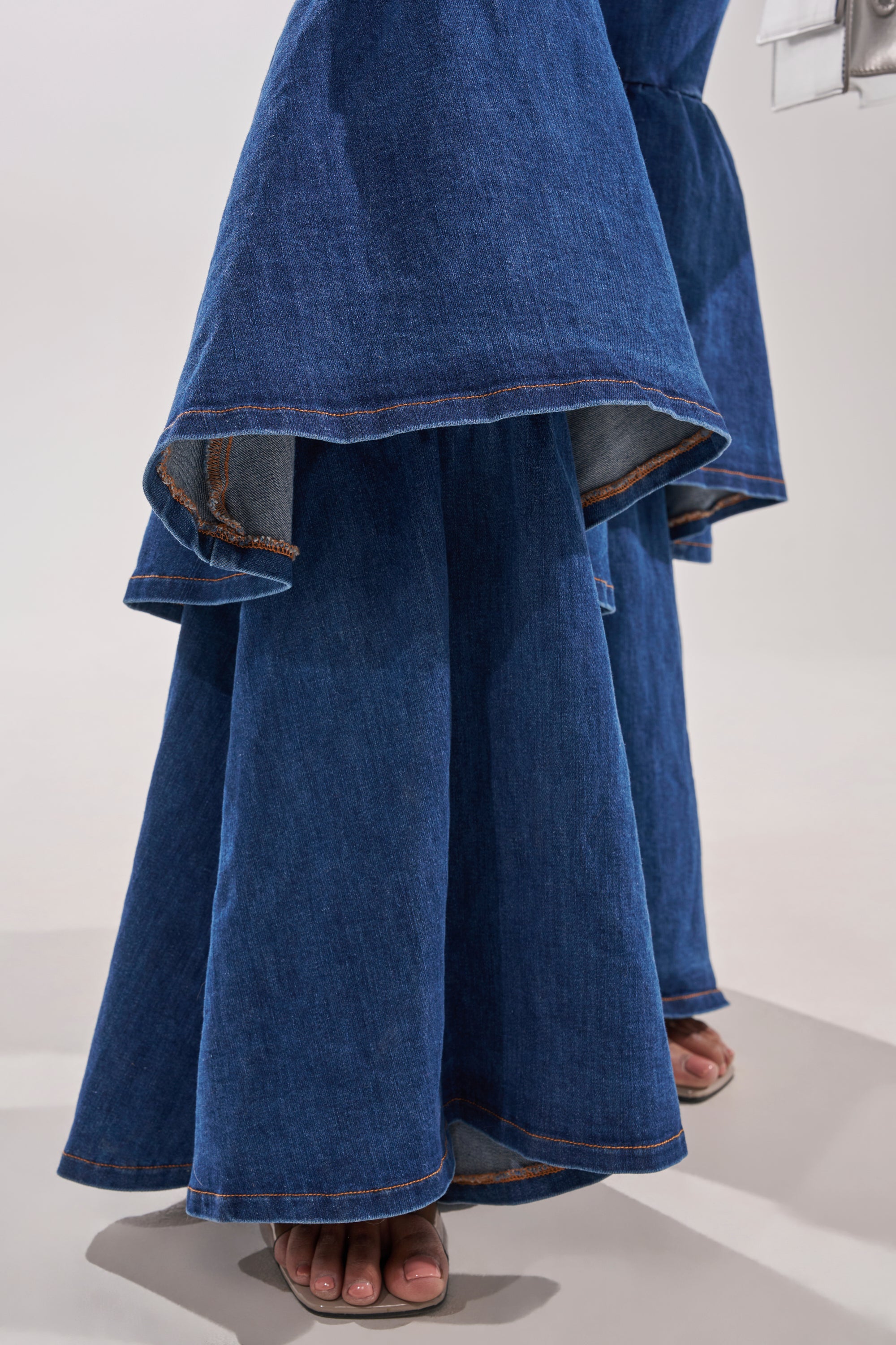 A close-up shows only the feet and lower legs of a person wearing the RODEO REBEL DENIM JUMPSUIT, featuring dramatic wide legs, bold layered hems, and heeled sandals for a retro-inspired statement look. 7