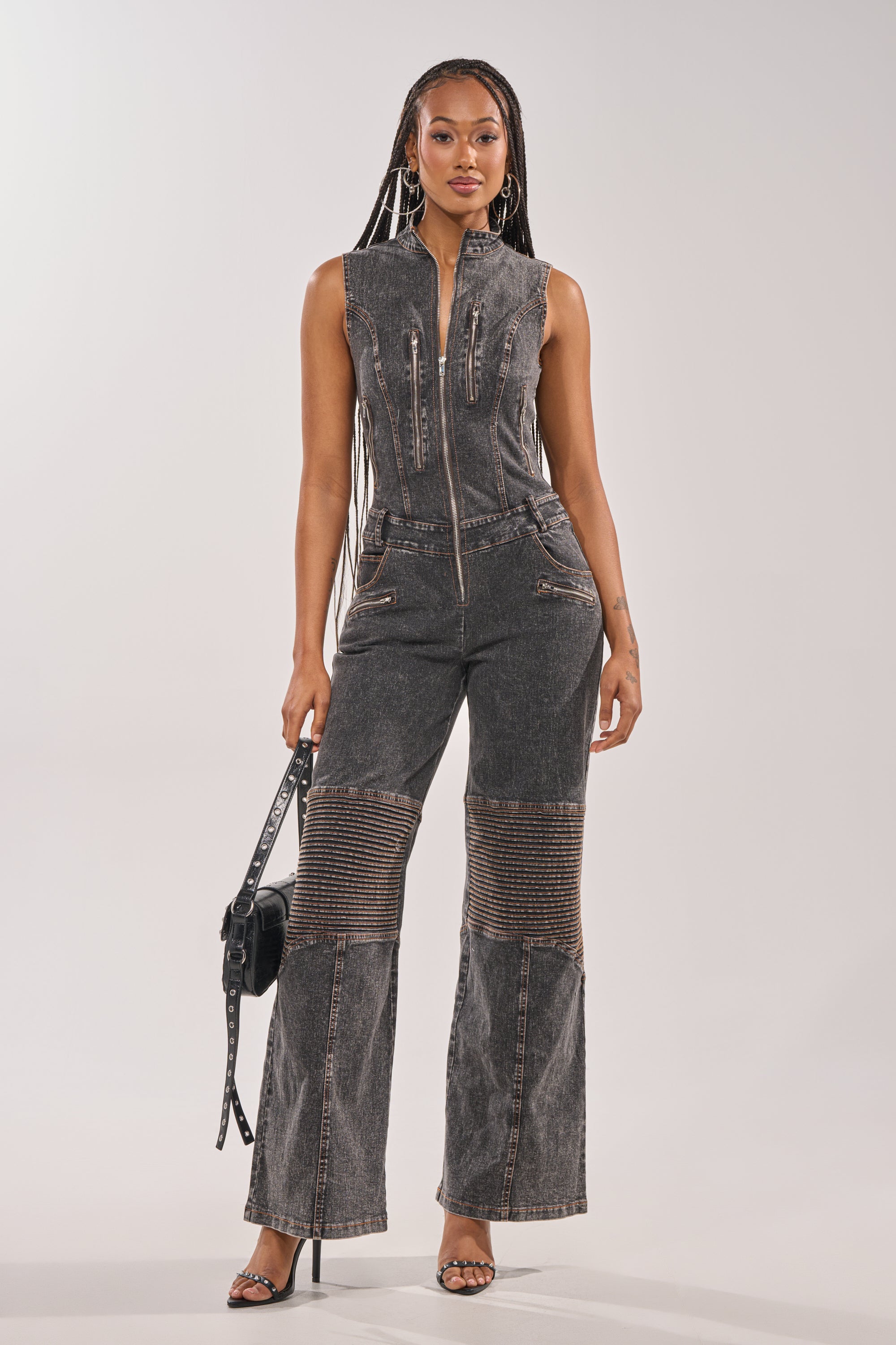 Radiating confidence, she wears the CAMILLA SLEEVELESS DENIM JUMPSUIT with moto-style knee stitching, paired with a black handbag, high-heeled sandals, long braids, and hoop earrings for a chic streetwear vibe. 0