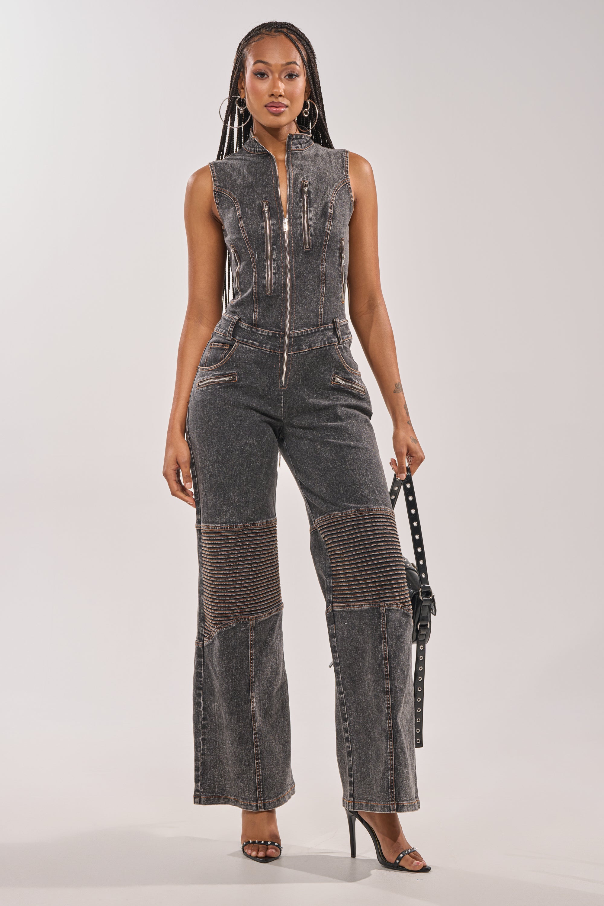 A woman stands confidently against a plain background in the CAMILLA SLEEVELESS DENIM JUMPSUIT, featuring moto-style textured leg panels, styled with black high-heeled sandals and holding a belt in one hand. 1