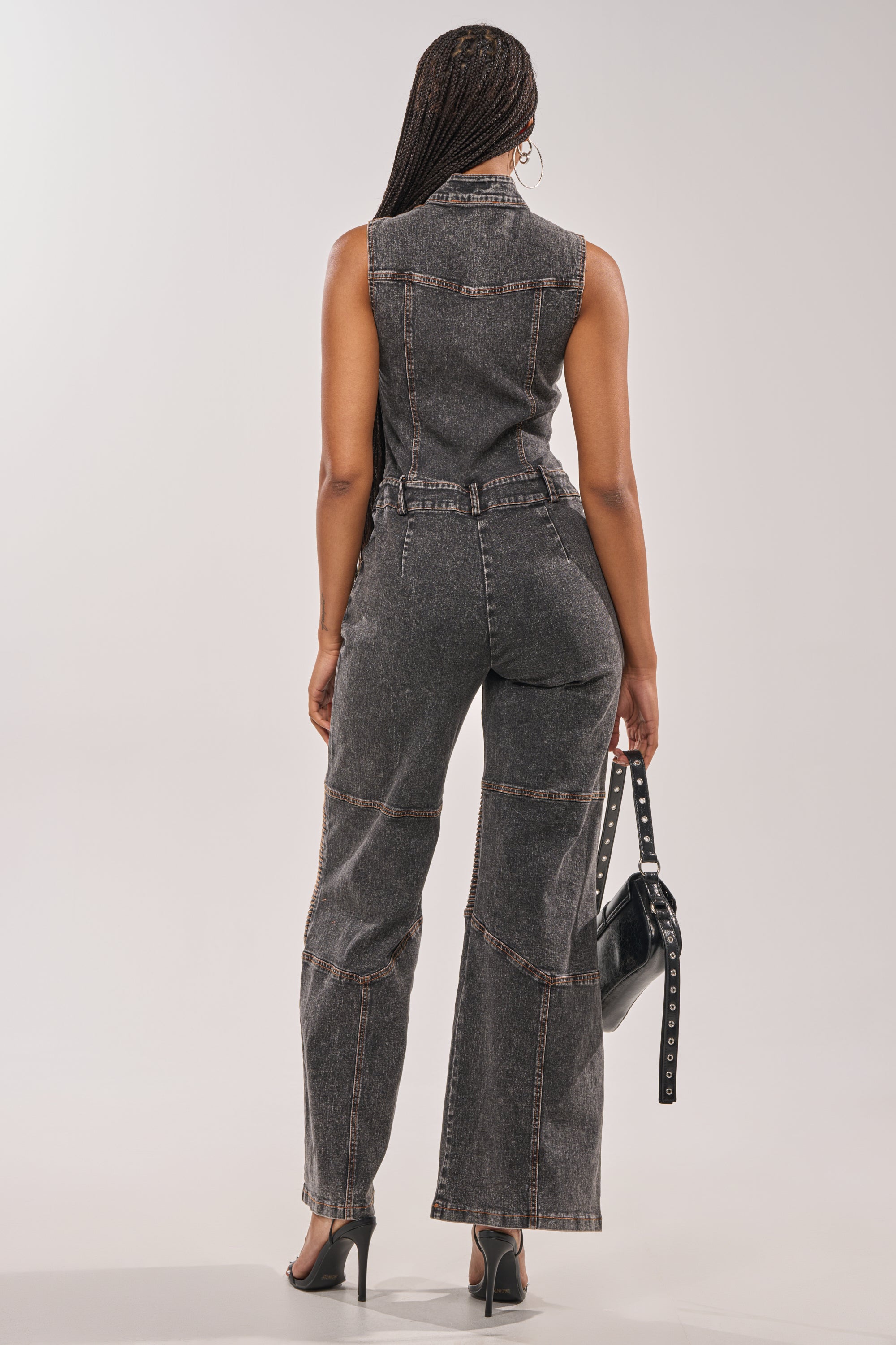 A woman with braided hair faces away, wearing the CAMILLA SLEEVELESS DENIM JUMPSUIT—dark gray, wide-leg with visible seams—paired with black high-heeled sandals and a black handbag, set against a neutral background. 3