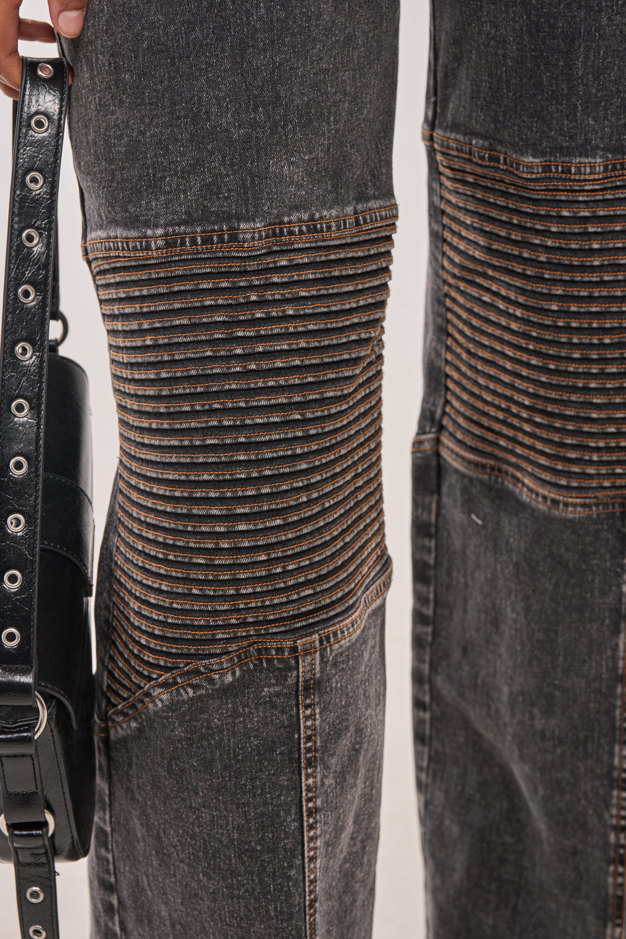 Close-up of the CAMILLA SLEEVELESS DENIM JUMPSUIT, featuring moto-style ribbed knee stitching. A person holds a black leather belt with metal holes beside their leg, giving this streetwear look an edgy touch. 5