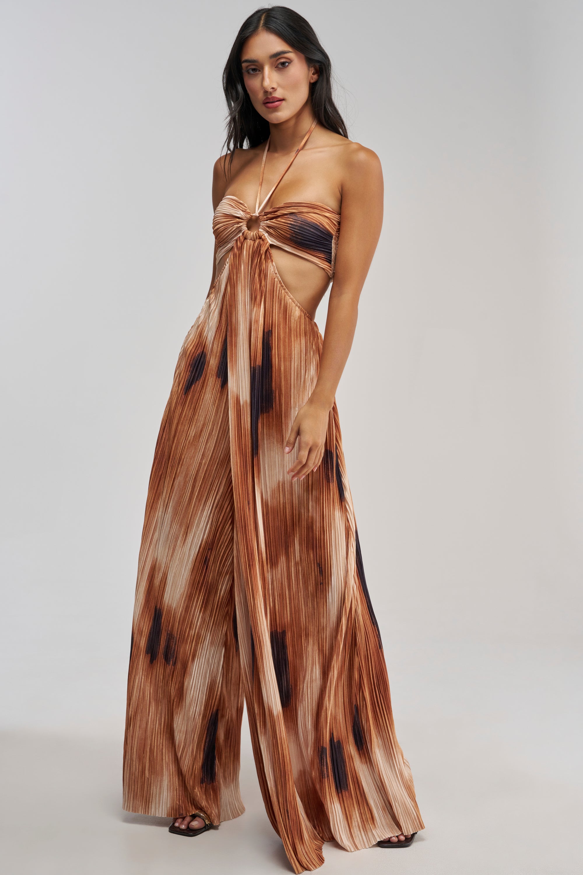 Wearing the FOUL PLAY PRINTED JUMPSUIT IN RUST—a pleated, brown and beige tie-dye satin halter jumpsuit with cut-out sides and wide legs—a woman stands against a plain light background, effortlessly showcasing summer style.