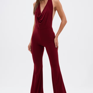A woman wears the LIKE MAGIC JUMPSUIT IN BURGUNDY, featuring a draped neckline and flared legs, styled with gold platform heels against a plain white background.