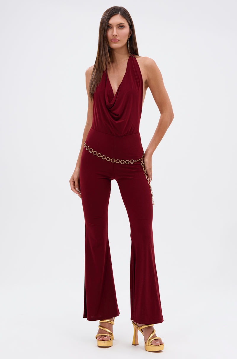 Woman wears the LIKE MAGIC JUMPSUIT IN BURGUNDY, a sleeveless cowl neck piece with flared legs, styled with a gold chain belt and platform heels, standing against a plain white background.