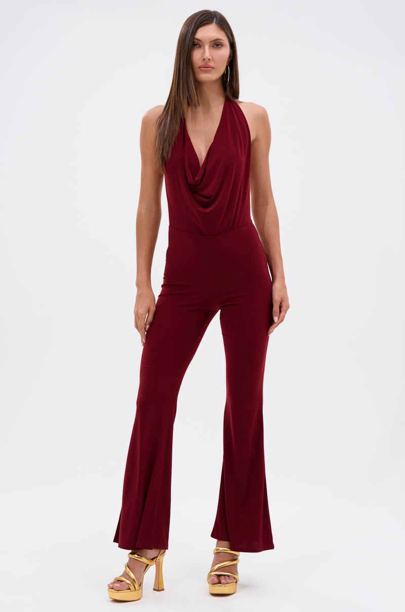 A woman faces forward in the LIKE MAGIC JUMPSUIT IN BURGUNDY, featuring a sleeveless halter-neck, draped neckline, and flared legs, styled with gold platform heels against a plain white background.
