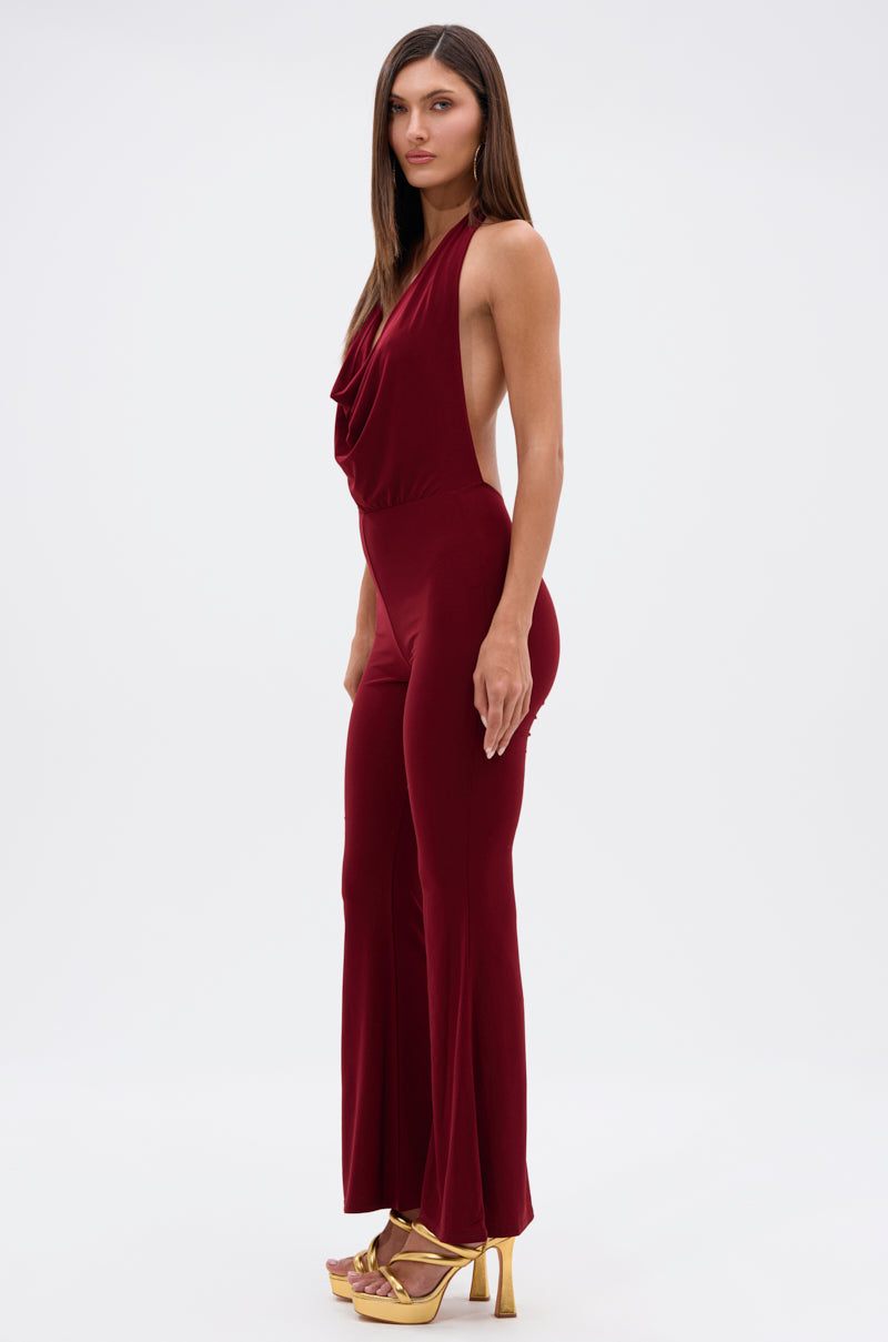A woman in profile wears the LIKE MAGIC JUMPSUIT IN BURGUNDY, featuring a sleeveless design, draped neckline, and open sides. She has long straight hair and gold high heels, posed against a white background.