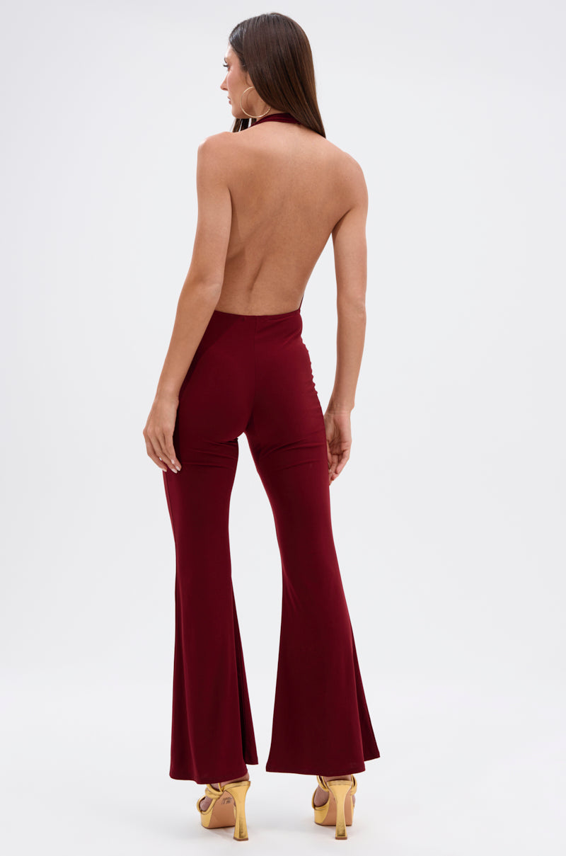 A woman faces away against a white background, wearing the LIKE MAGIC JUMPSUIT IN BURGUNDY—a halter-style jumpsuit with flared legs—paired with yellow high heels.