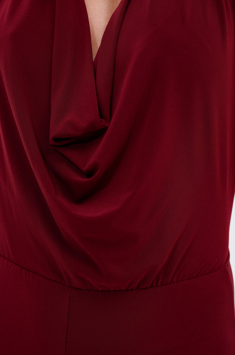 Close-up of a person wearing the LIKE MAGIC JUMPSUIT IN BURGUNDY, featuring a draped cowl-neck. The image spotlights the neckline and upper torso, highlighting its smooth fabric, elegant folds, and rich burgundy hue.